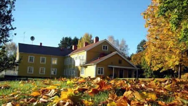 Une maison jaune à deux étages, avec un toit rouge, entourée de feuilles d’automne.