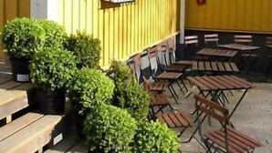 Un bâtiment jaune avec une fenêtre et une porte, des tables et des chaises à l’extérieur pour créer un espace de détente, et des plantes en pot.