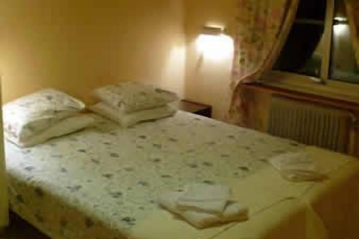 Ein Einzelbett mit floralem Überwurf und Kissen, ein Nachttisch mit Lampe und ein Fenster mit Vorhängen.