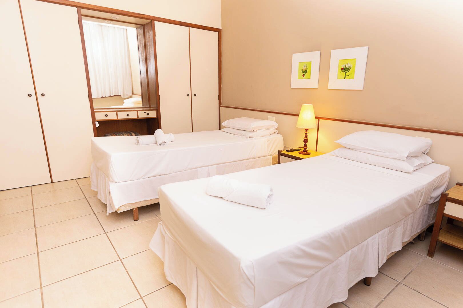 Een hotelkamer met twee eenpersoonsbedden, een garderobe, een spiegel, een nachtkastje met een lamp, en twee ingelijste foto’s aan de muur.
