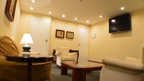 A hotel room with a sofa, armchair, and a small table. There are two doors, a flat-screen TV mounted on the wall, and a lamp on a side table. - Rede Andrade Hotel Canadá (Rio de Janeiro)