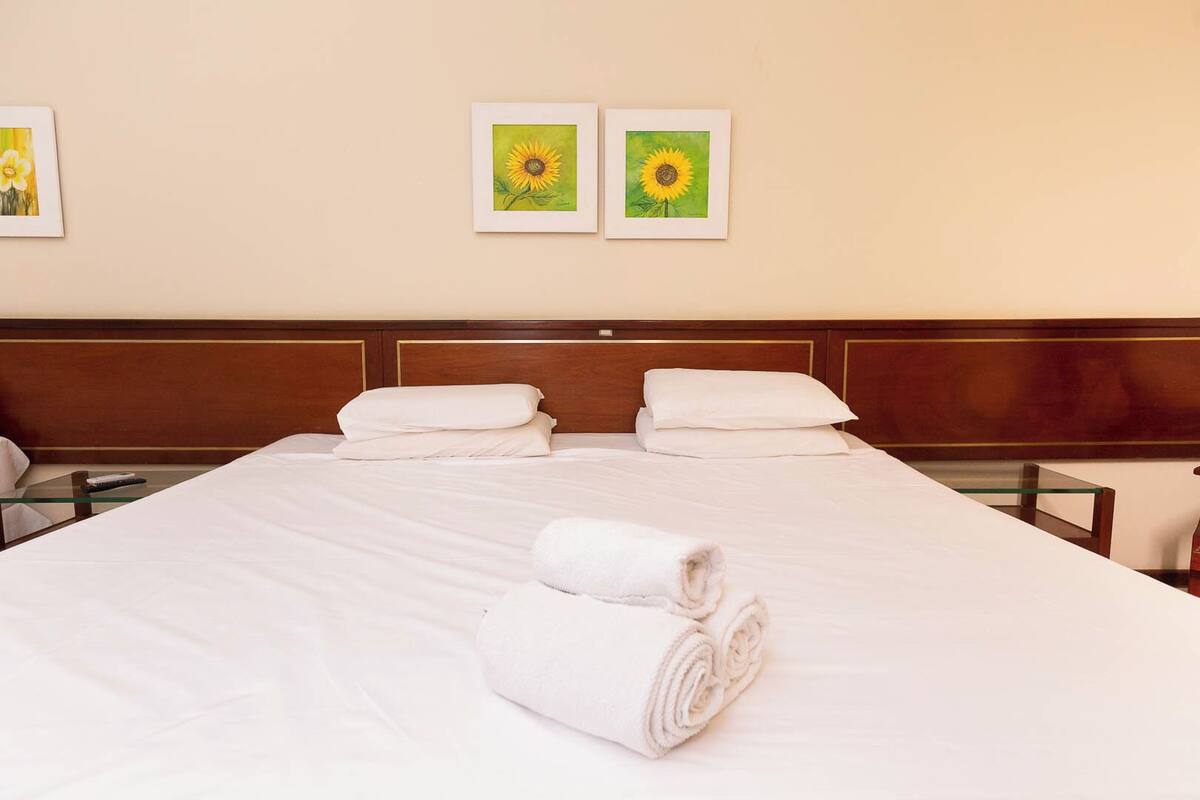 Una cama con arreglos impecables, sábanas blancas y dos toallas enrolladas, un cabecero de madera y dos girasoles enmarcados en la pared.