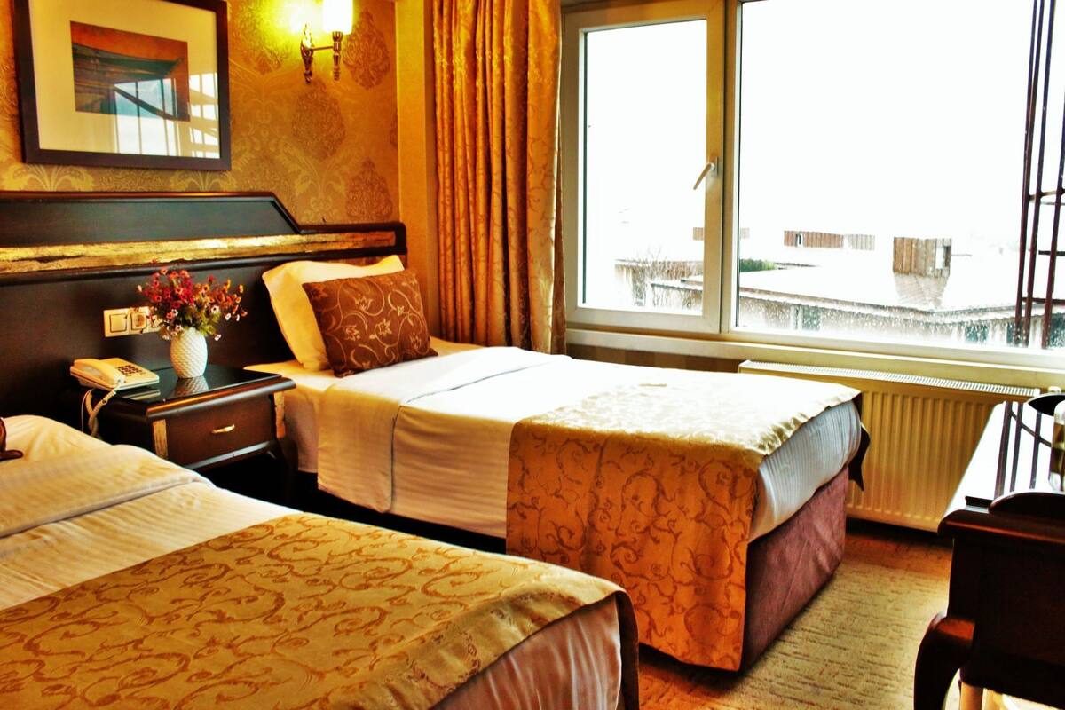 A hotel room with two beds, a bedside table with a phone, a vase with flowers, and a window with a view of buildings.