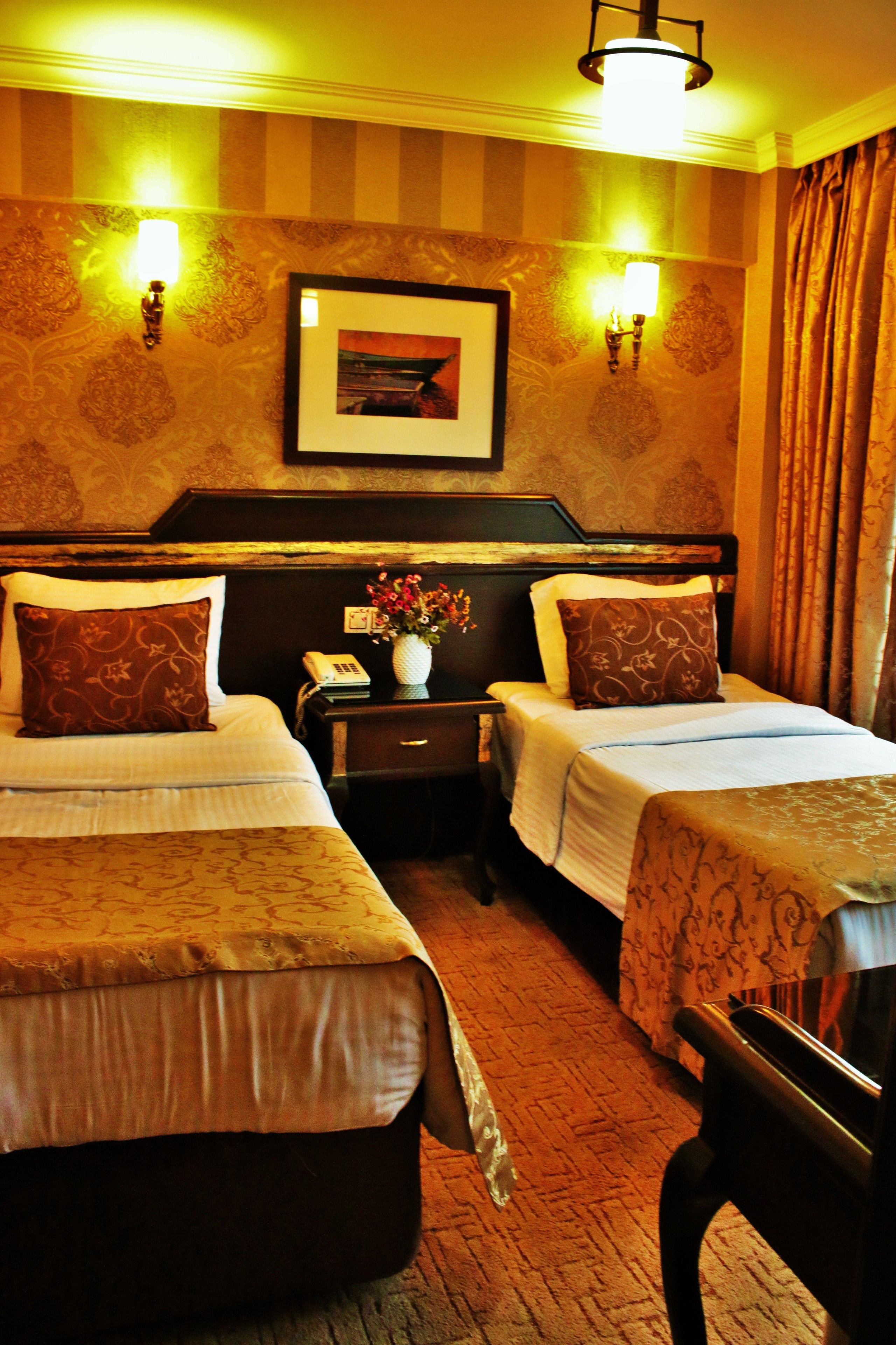 A hotel room with two beds, a nightstand with a telephone and a vase of flowers, and a framed picture on the wall.