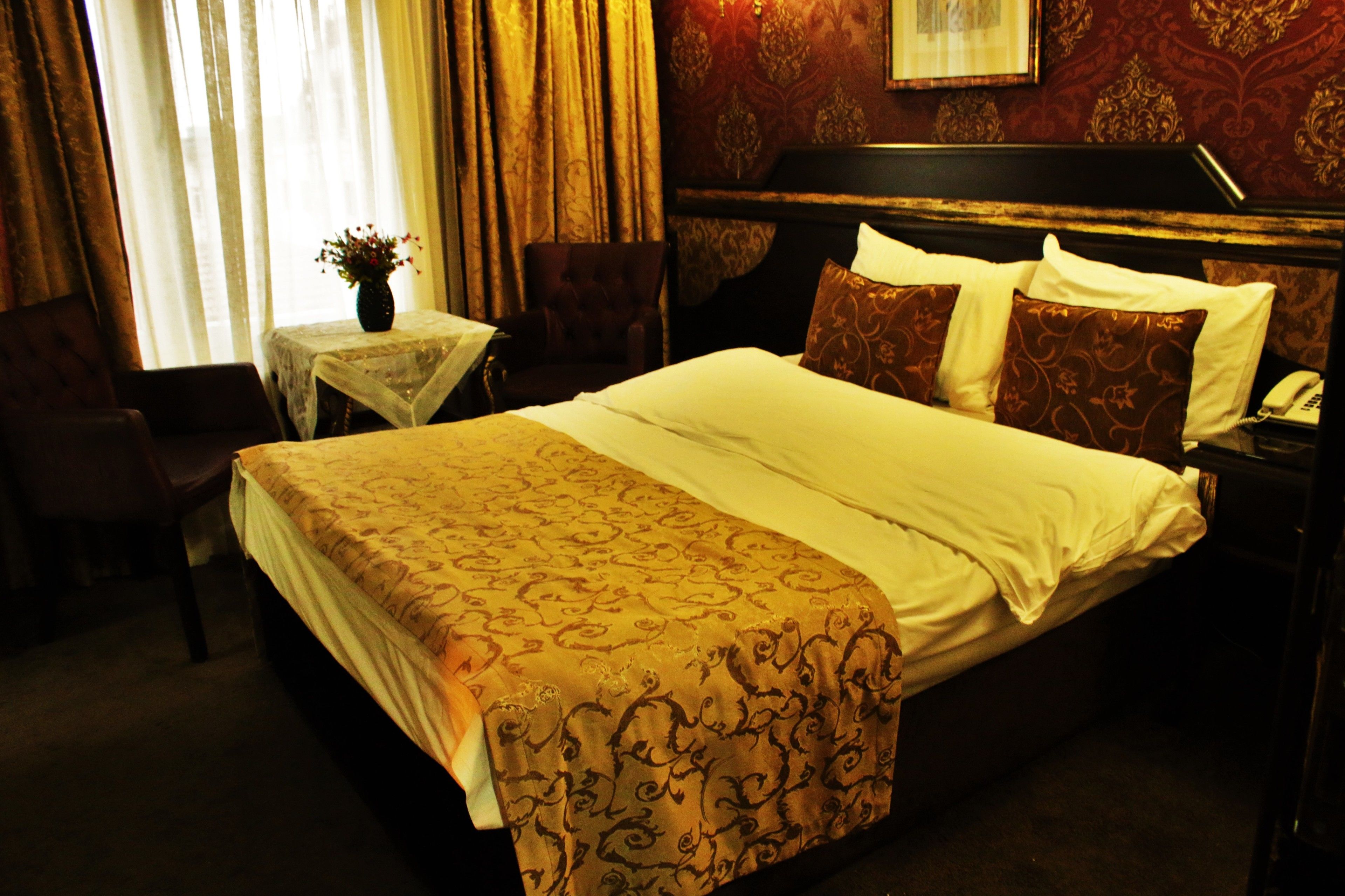 Habitación de hotel con una cama grande, una mesita de noche, una silla, una pequeña mesa con un florero y una ventana con cortinas.