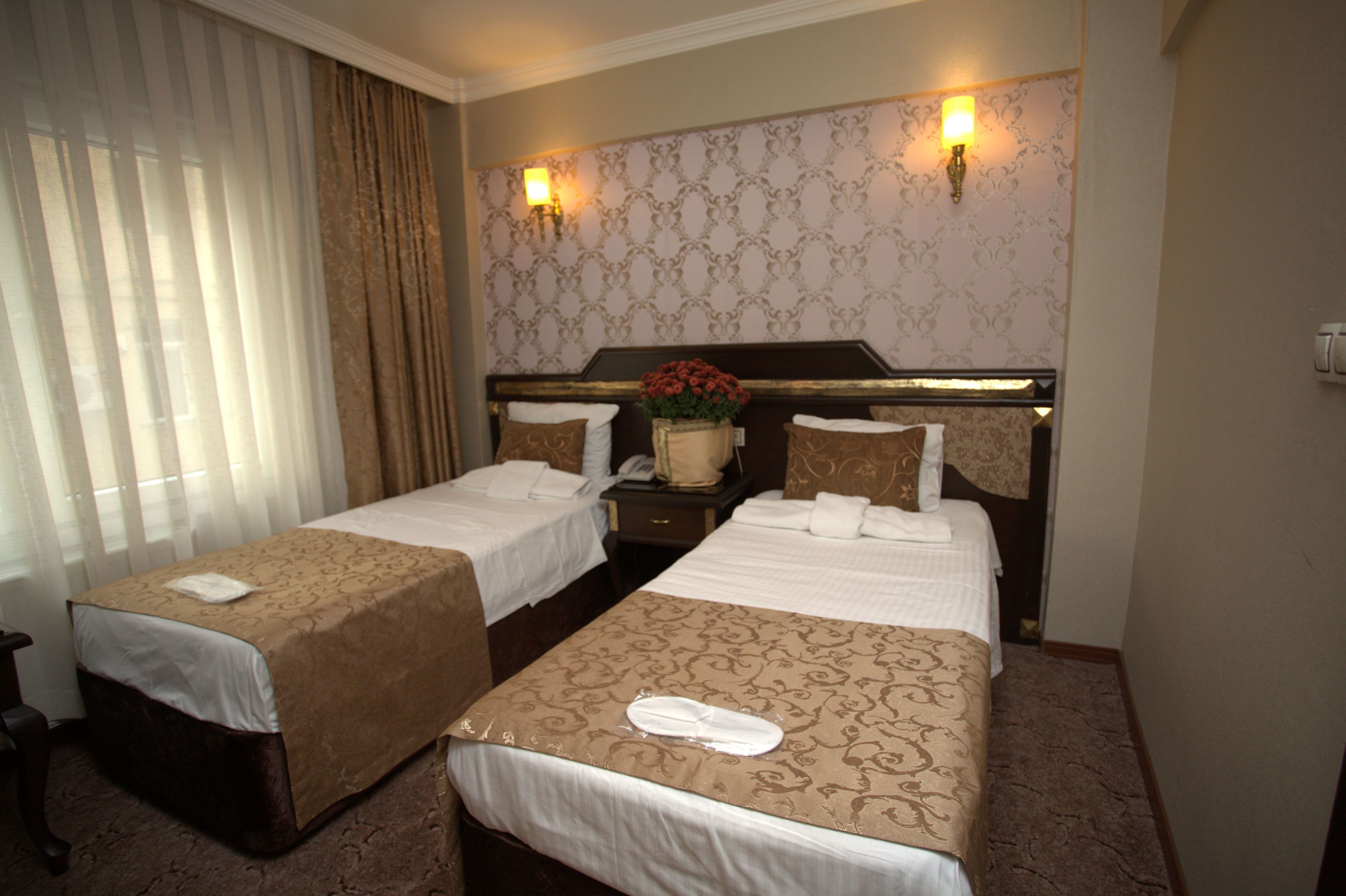 A hotel room with two beds, a nightstand, a vase with flowers, and a wall-mounted light fixture.