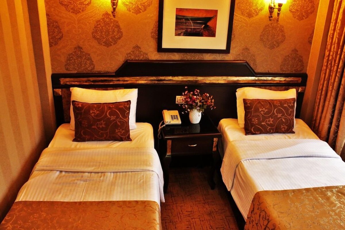 Habitación de hotel con dos camas, una mesita de noche con teléfono y un florero, y un cuadro en la pared.