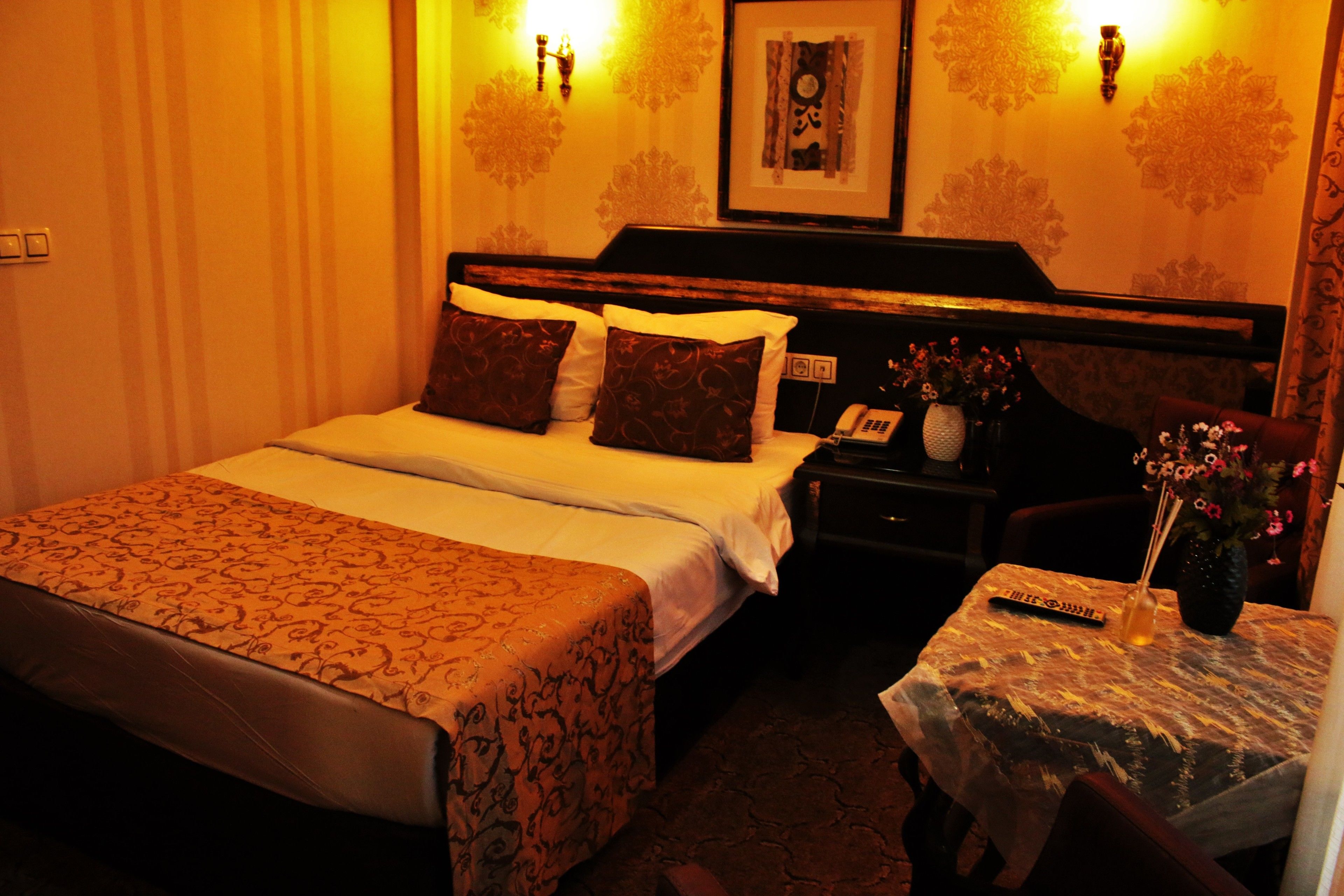A hotel room with a bed, bedside table, and a small table with a vase of flowers.