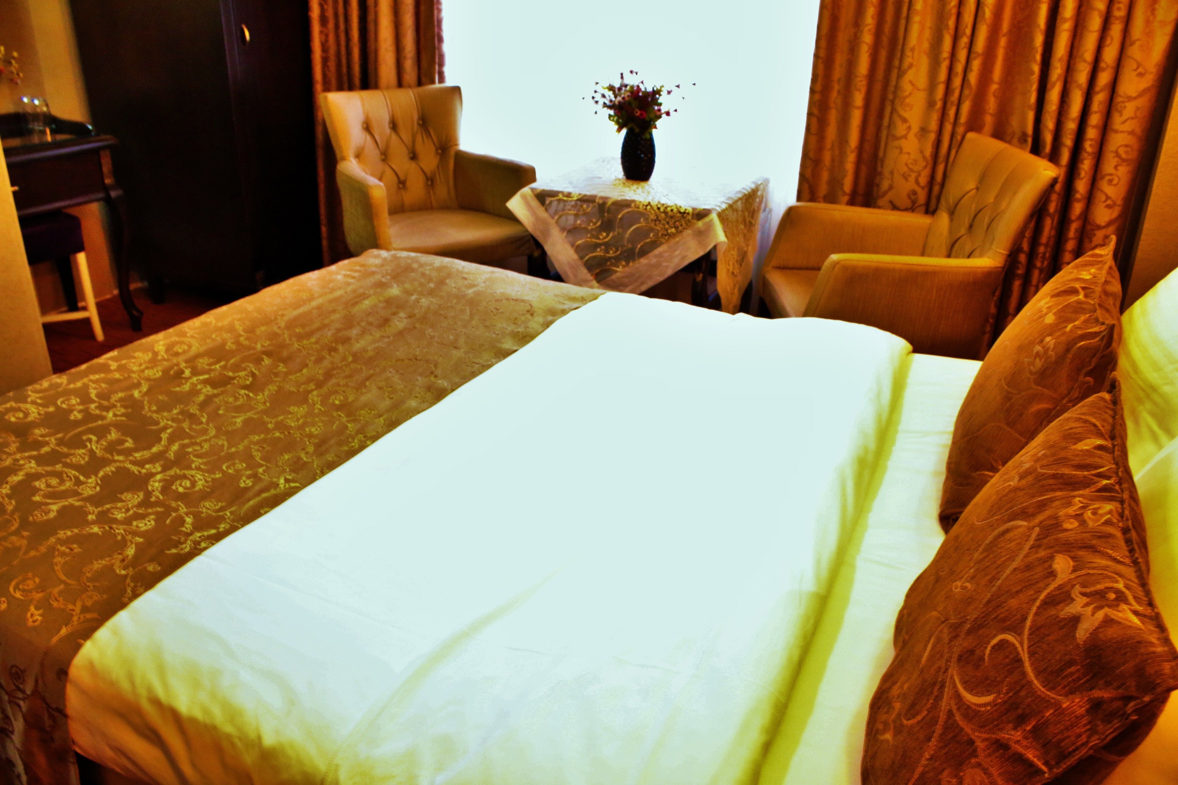 Una habitación de hotel con una cama grande, dos sillones, una mesita con un florero y cortinas.