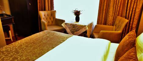 Une chambre d’hôtel avec un grand lit, deux fauteuils, une petite table avec un vase et des rideaux.