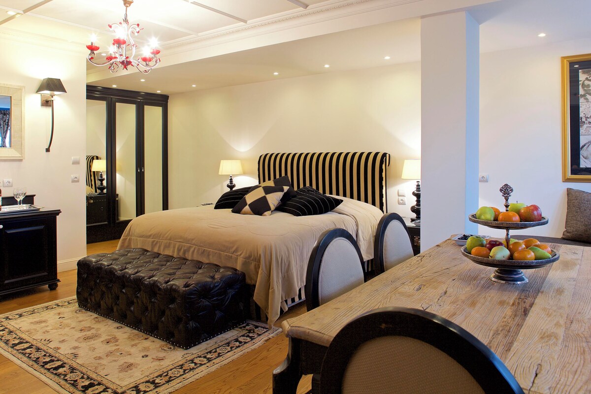 A bedroom with a large bed, a wooden dining table with fruit, a black leather ottoman, and a chandelier.