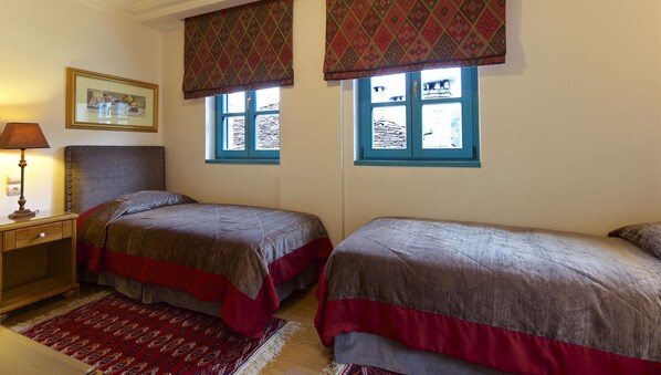 Una habitación con dos camas, una mesita de noche con lámpara, un tapete estampado y dos ventanas con cortinas decoradas.