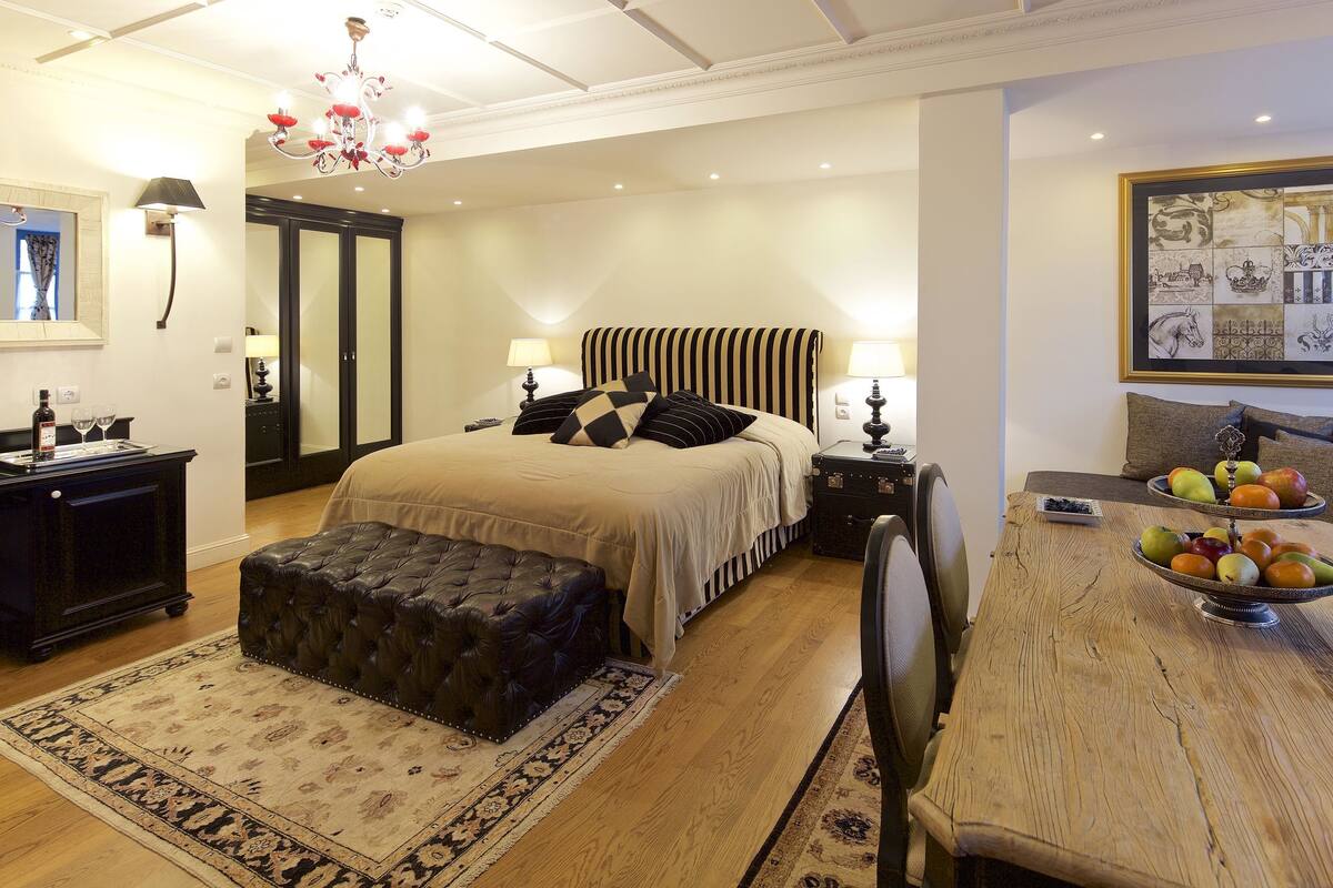 Een slaapkamer met een groot bed, een houten eettafel met fruit, een leren voetenbank en een kroonluchter.