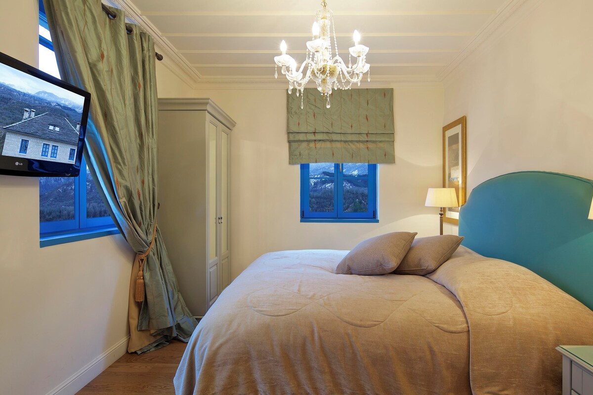 A bedroom with a bed, a chandelier, a window with a view, a bedside table, and a lamp.
