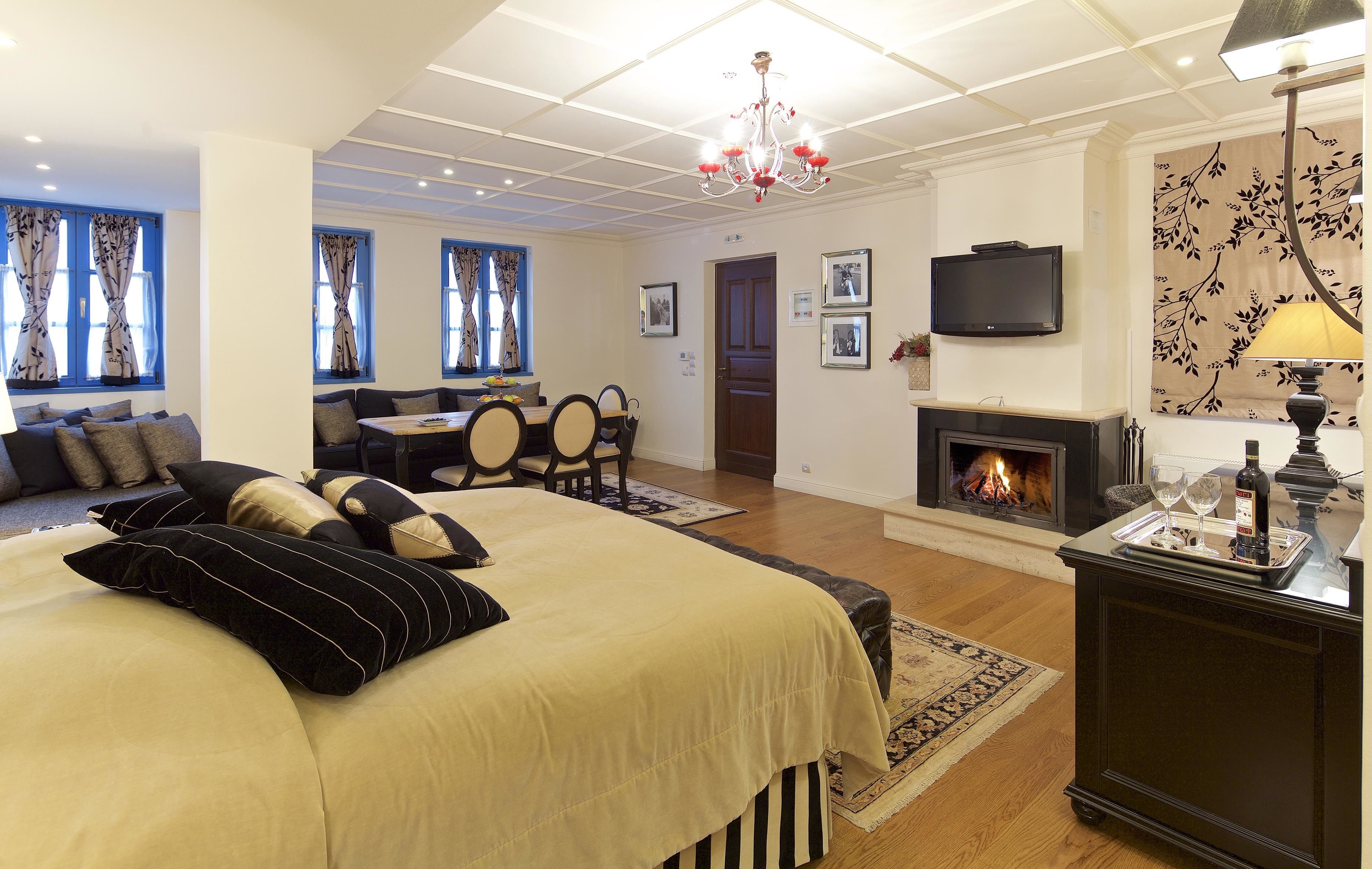 A hotel room with a large bed, a sofa, a dining table, chairs, a fireplace, a TV, and a chandelier.