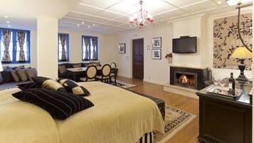 A hotel room with a large bed, a sofa, a dining table, chairs, a fireplace, a TV, and a chandelier.