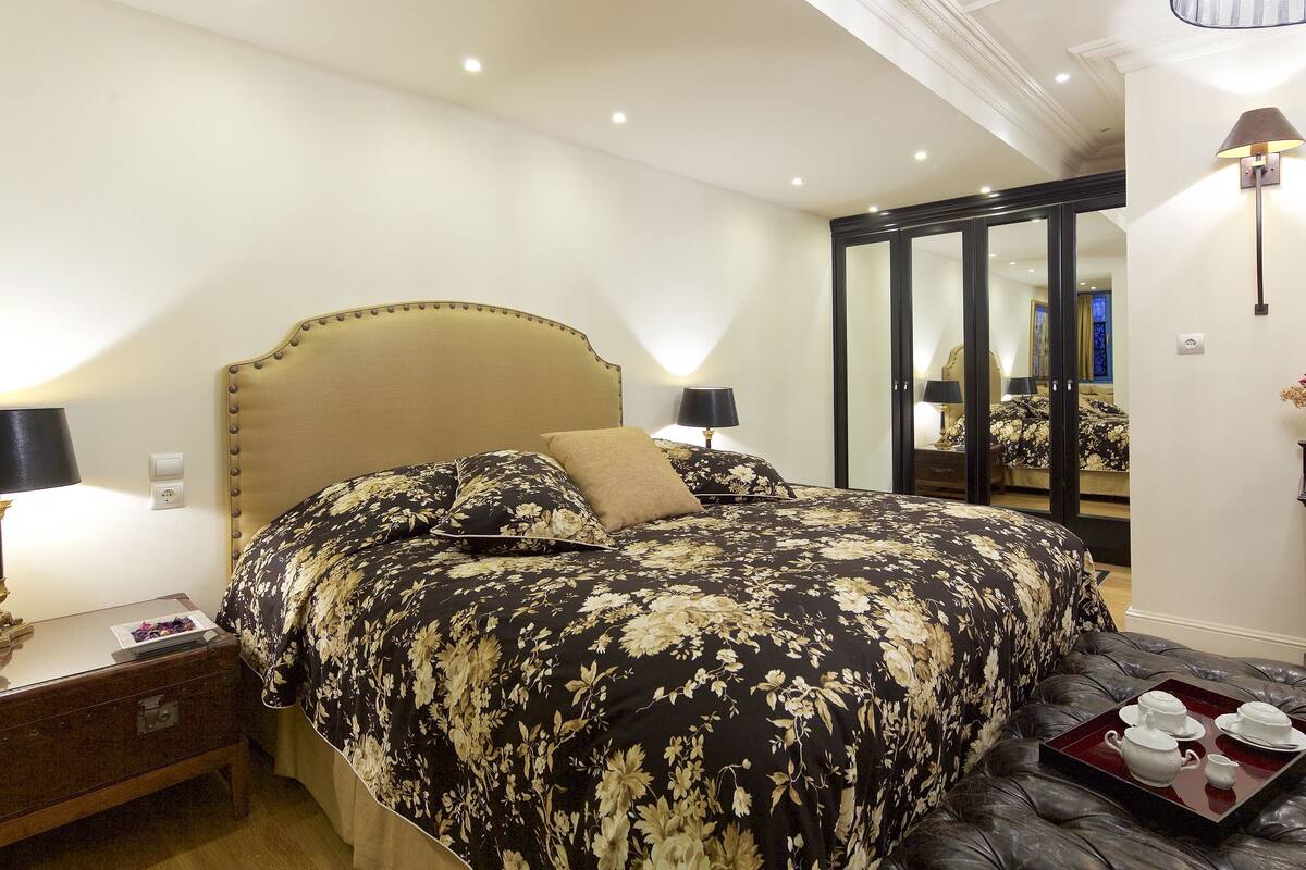 Een slaapkamer met een bed, nachtlampjes, een nachtkastje, een kaptafel en een dienblad met theekopjes.