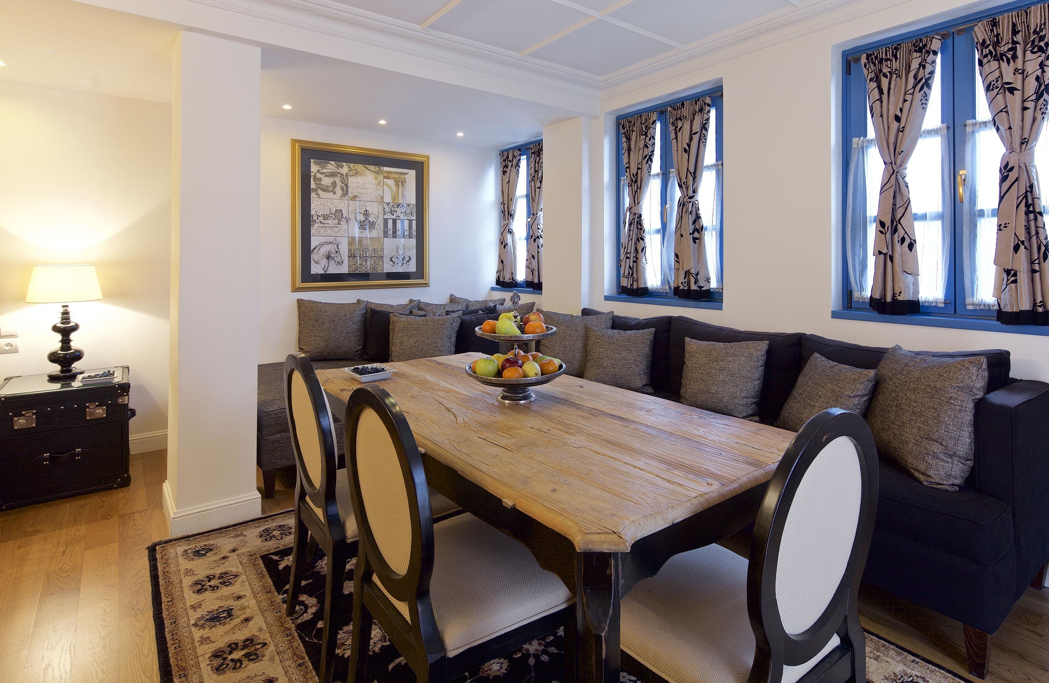Un comedor con una mesa de madera, sillas y un bol de fruta. Se ve un sofá y una lámpara al fondo.
