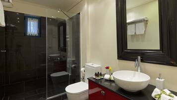 Un baño moderno con ducha envuelta en vidrio, inodoro blanco, tocador rojo con un lavabo grande y blanco, y un espejo sobre el lavabo.