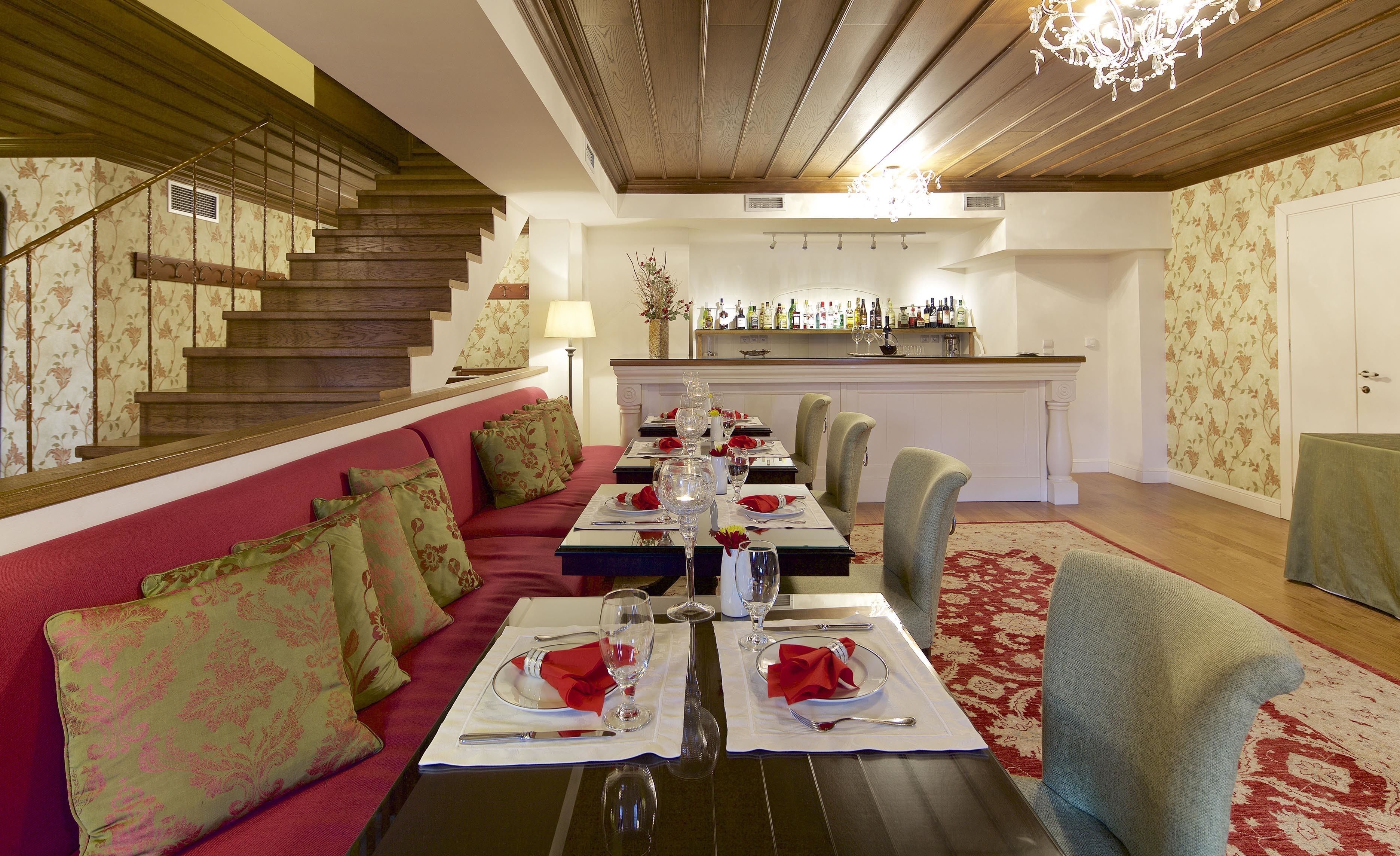 A dining area with a long table set for six, a red sofa with patterned cushions, a staircase with wooden railings, and a bar area with a wine display.