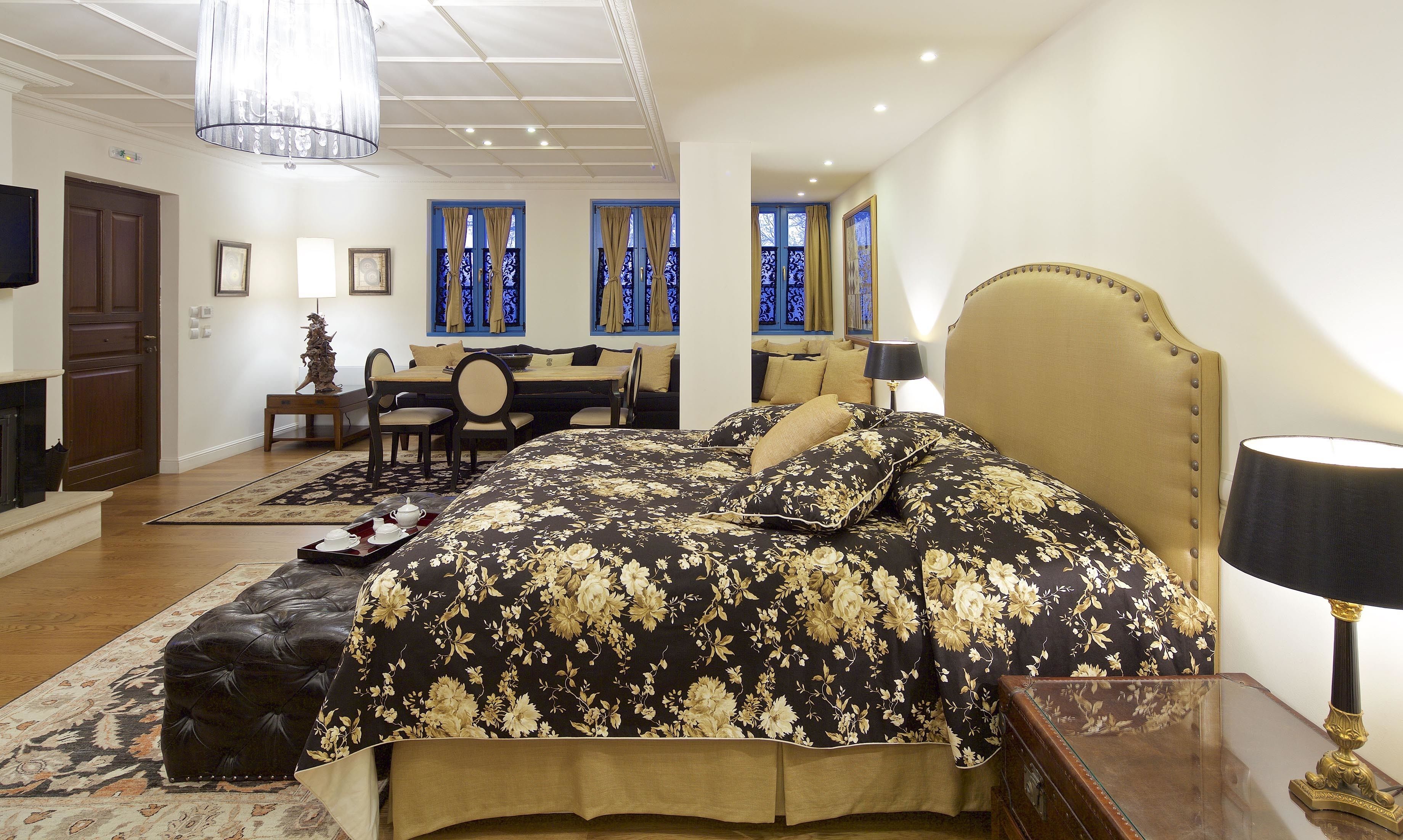 A bedroom with a large bed, a desk, and a chair. There is a fireplace, a chandelier, and a window with curtains.