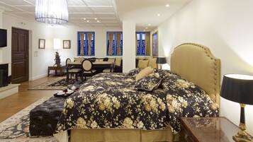 A bedroom with a large bed, a desk, and a chair. There is a fireplace, a chandelier, and a window with curtains.