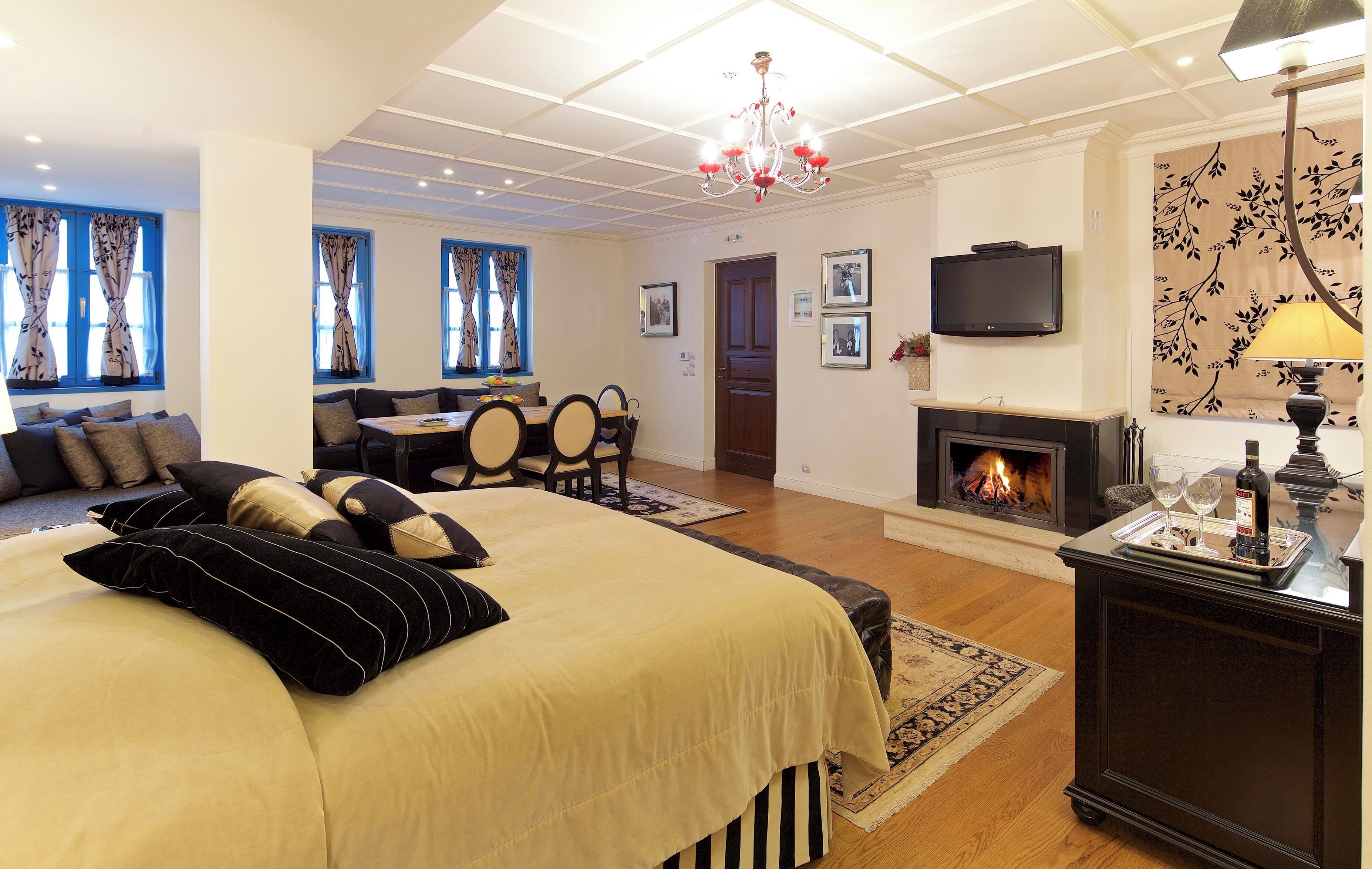 A hotel room with a large bed, a sofa, a dining table, chairs, a fireplace, a TV, and a chandelier.