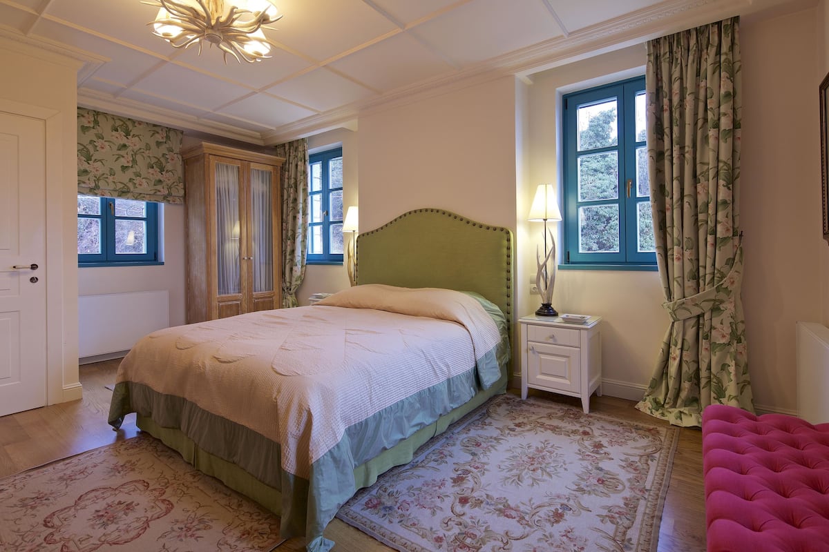 Een slaapkamer met een bed, nachtkastje, lamp, bloemen gordijnen en een roze stoel.