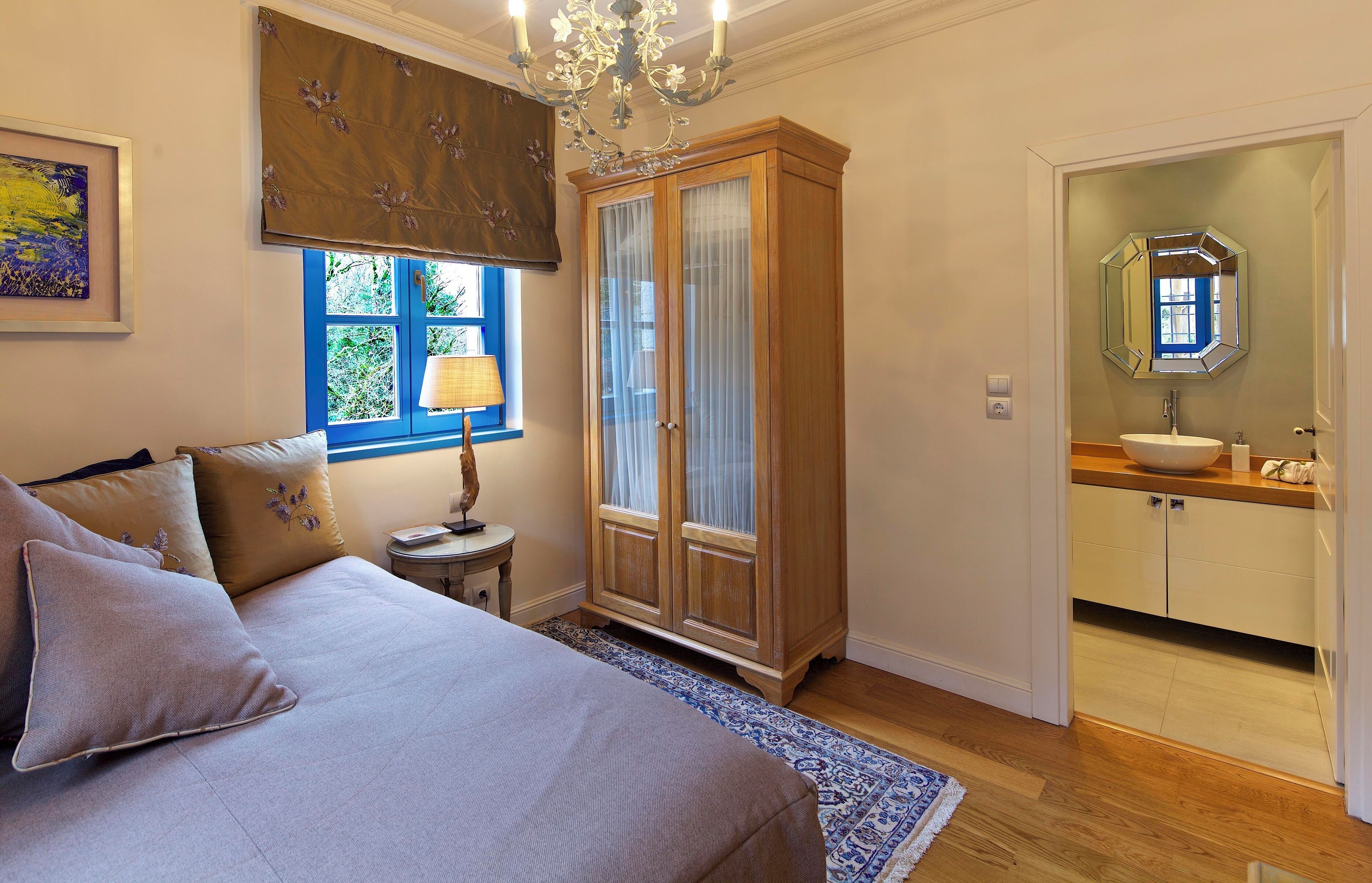 A bedroom with a bed, a nightstand, a window with curtains, a wardrobe, and a bathroom with a sink and mirror.