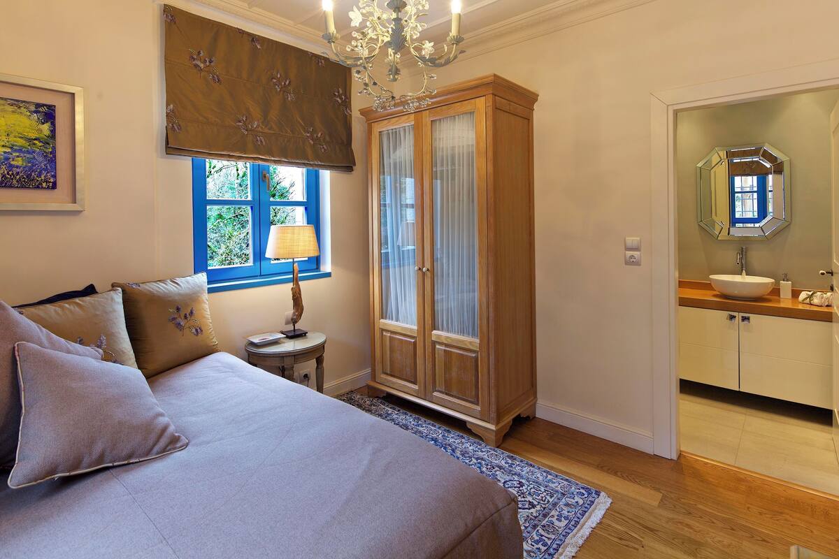 Een slaapkamer met een bed, nachtkastje, raam met gordijnen, kledingkast en een badkamer met wastafel en spiegel.