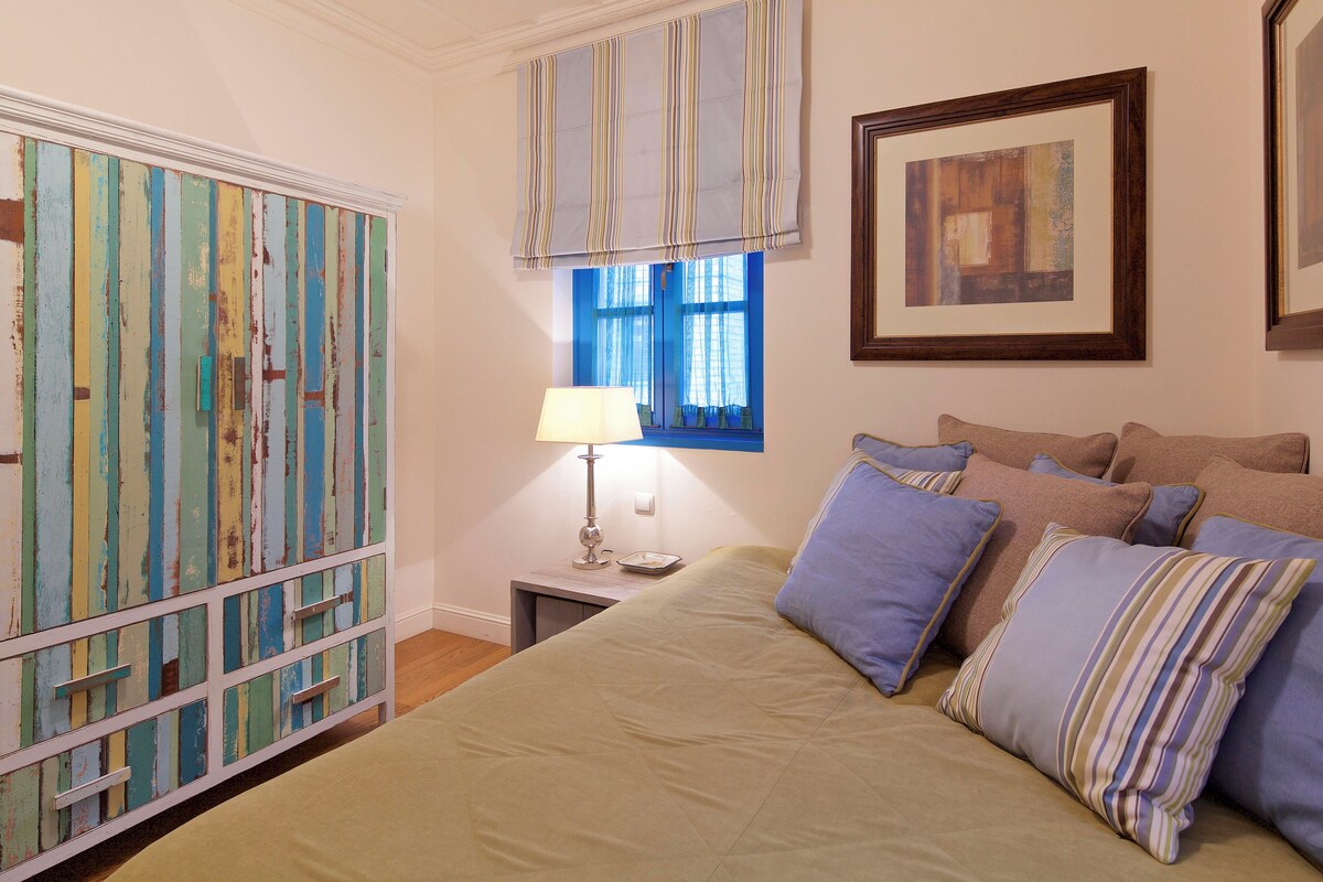 Een slaapkamer met een bed, nachtkastje, lamp, raam met gordijnen en een kunstwerk aan de muur.