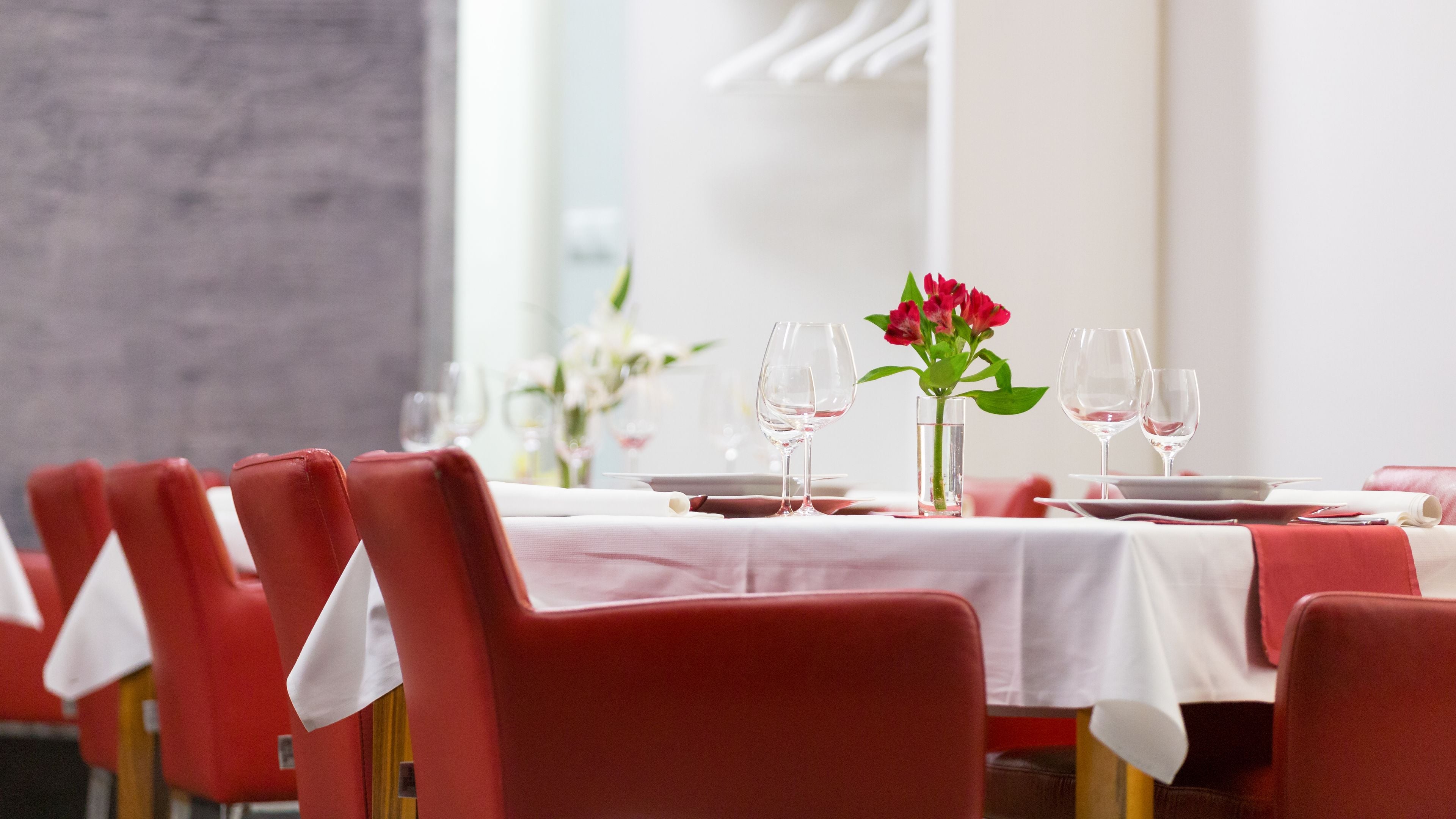 A dining area with red chairs, white tablecloths, and a vase of red flowers.
