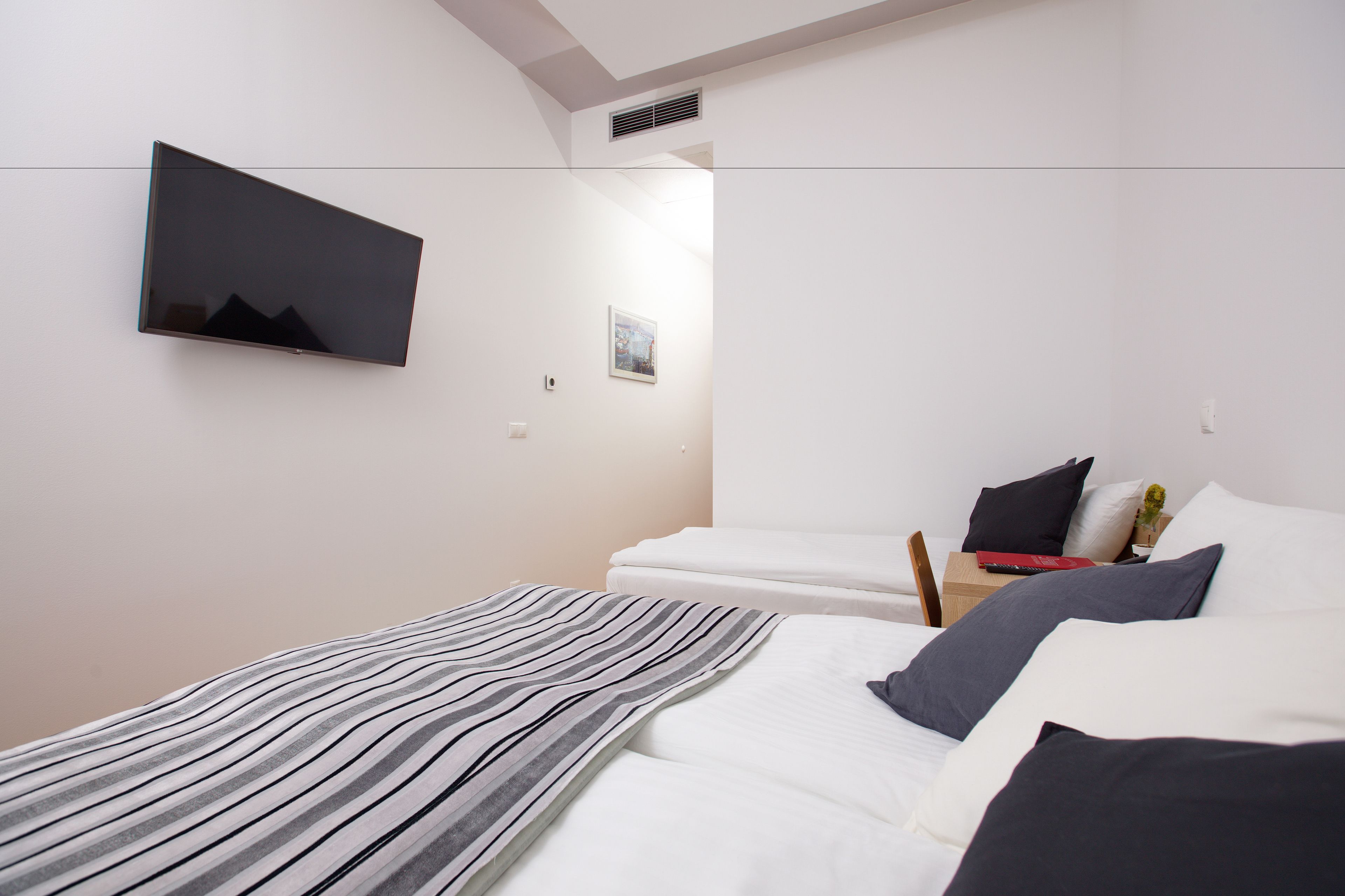 Una habitación de hotel moderna con un televisor de pantalla plana en la pared, una cama bien hecha con ropa blanca, un plaid a rayas y una pequeña mesita de noche con una lámpara y un libro.