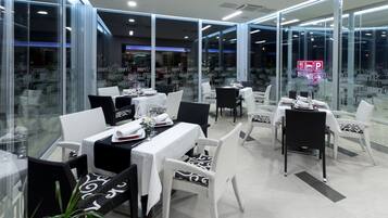 Een modern restaurant met glazen wanden, zwart-wit rieten meubels en netjes opgemaakte tafels met witte tafelkleden.