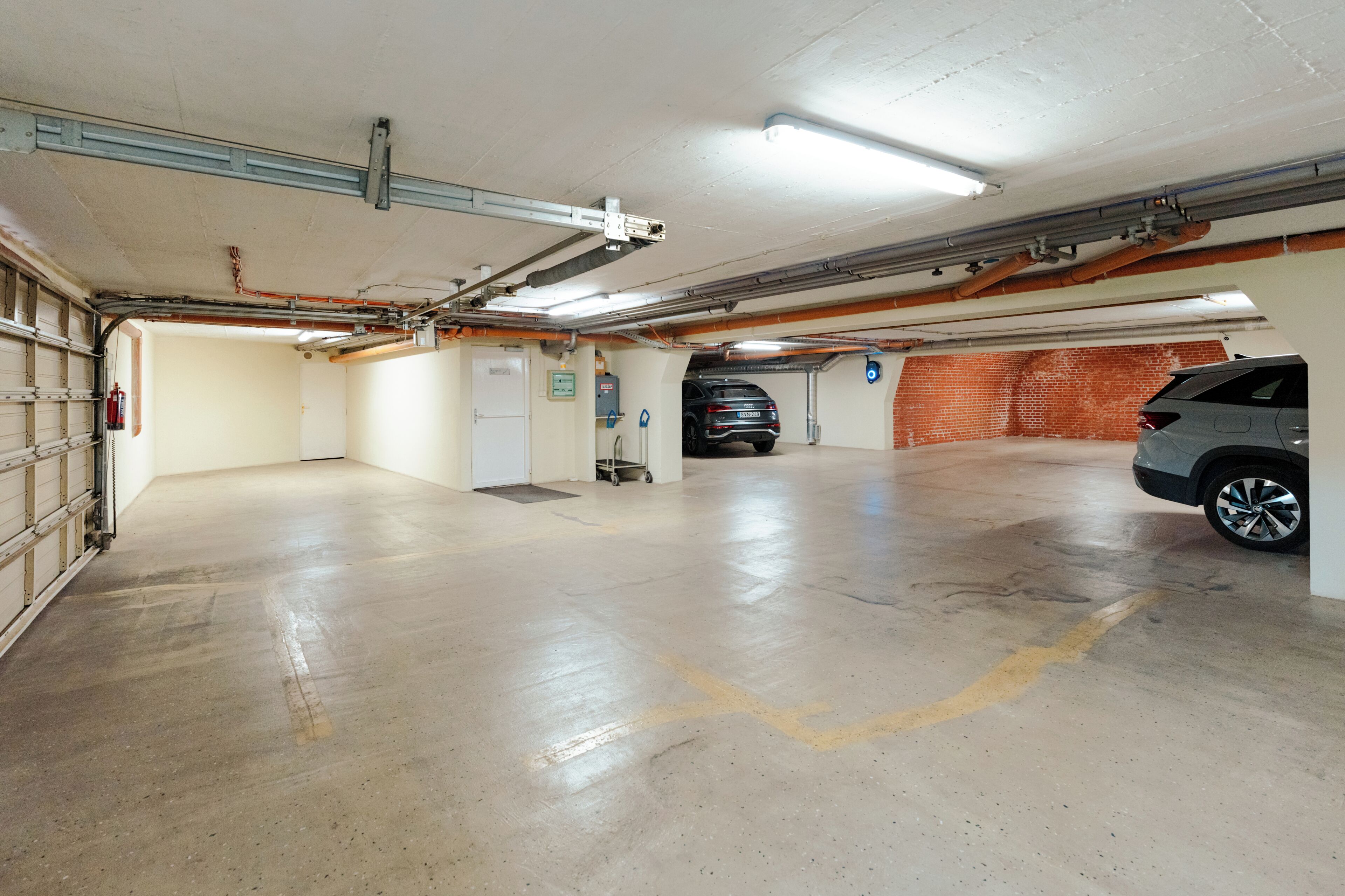 A parking garage with concrete floors, a fire extinguisher, a fire alarm, and a vehicle parked in the distance.
