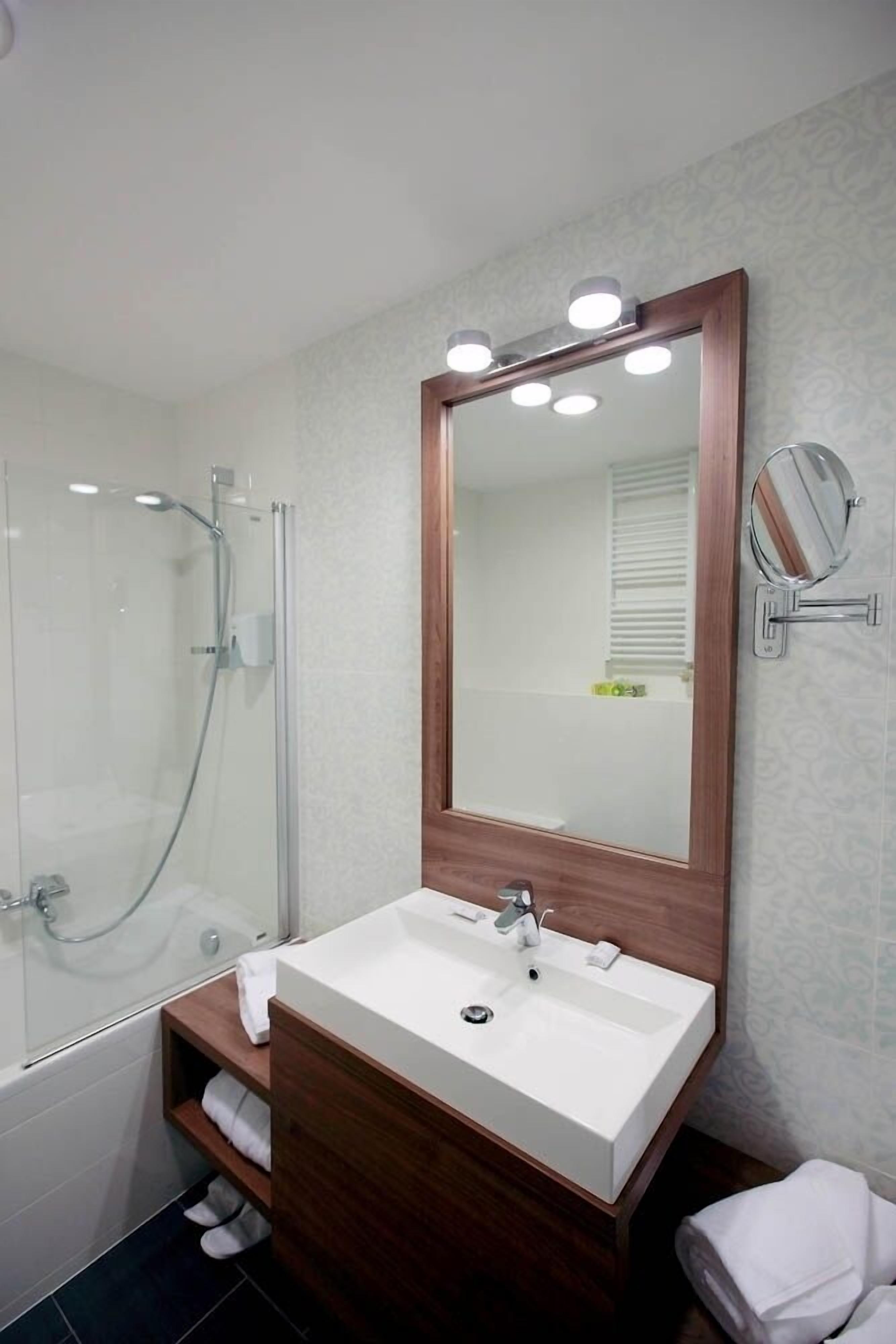 A modern bathroom with a glass-enclosed shower, a white sink with a wooden vanity, and a large mirror with lights.