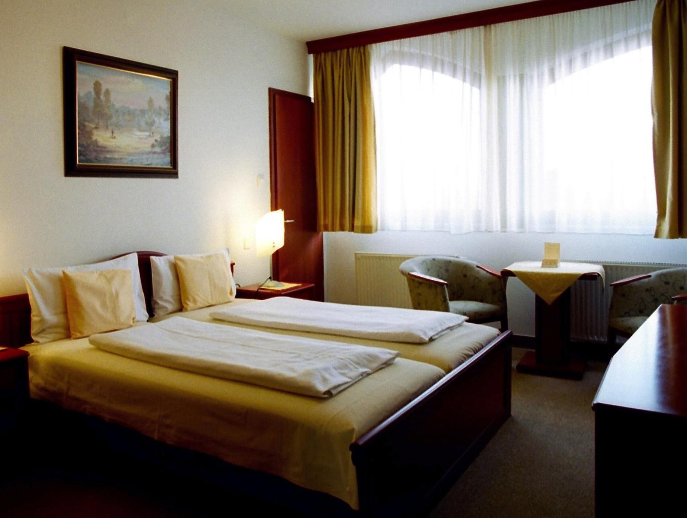 A hotel room with two beds, a desk, a chair, a painting on the wall, and a window with curtains.