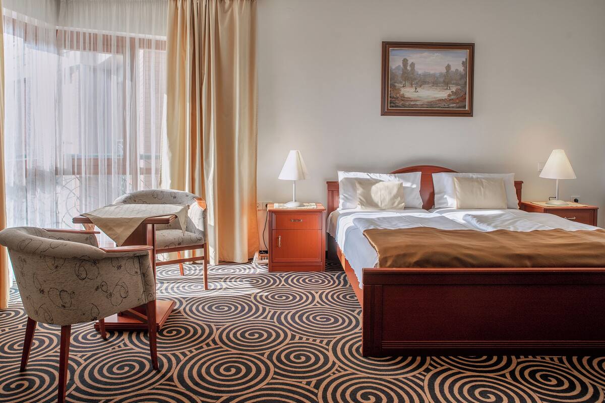 A hotel room with a bed, two bedside tables with lamps, a chair, a small table, and a large window with curtains.