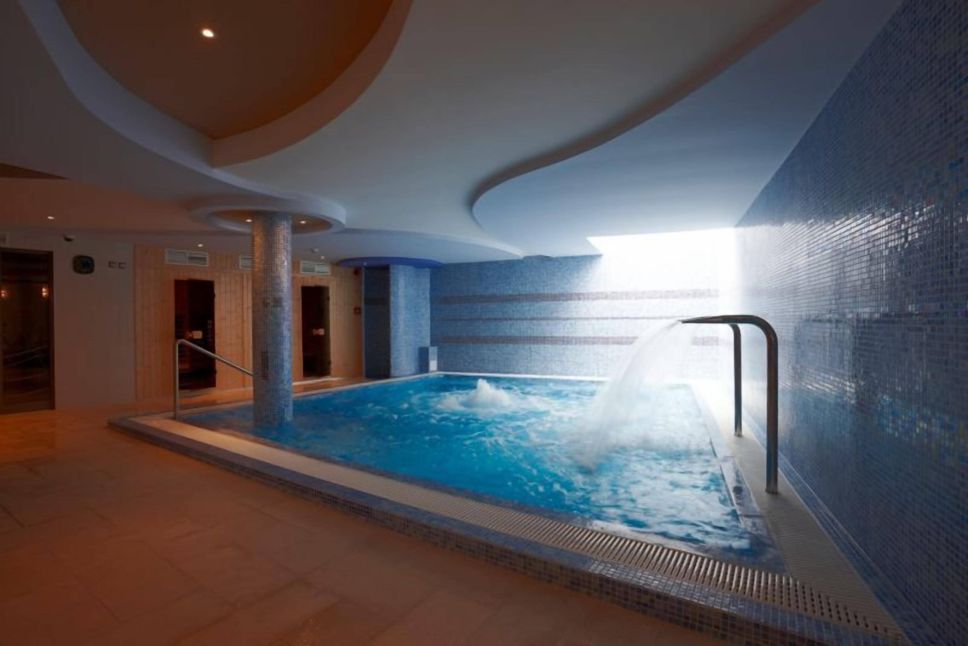 A large indoor swimming pool with a water jet feature, surrounded by a tiled wall and a curved ceiling.