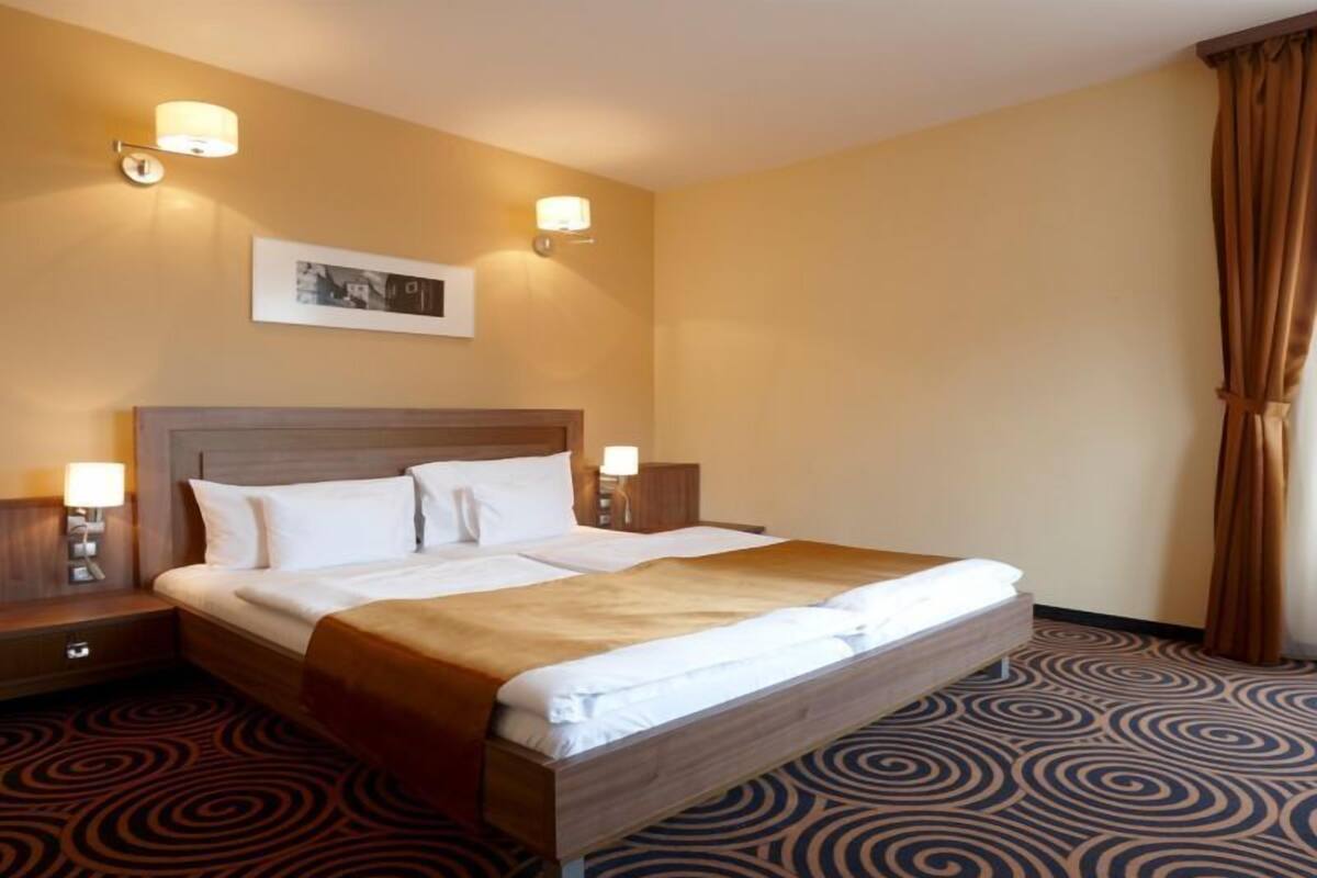 A hotel room with a large bed, two bedside tables with lamps, a headboard, a patterned carpet, and a framed picture on the wall.
