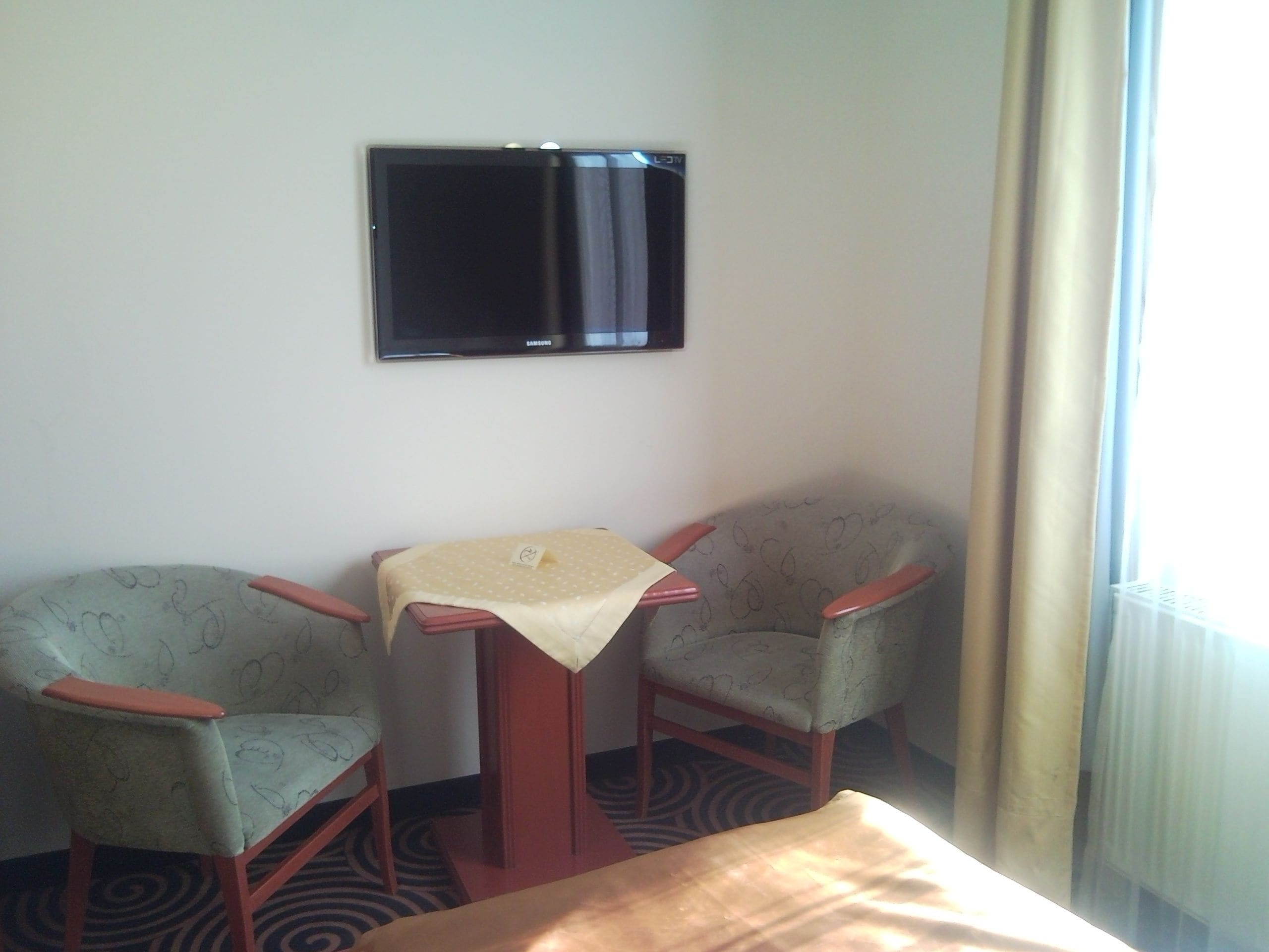Habitación de hotel con dos sillones, una mesita con mantel, un televisor de pantalla plana en la pared y una ventana con cortinas.