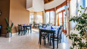 A spacious dining area with multiple tables and chairs, large windows with curtains, and potted plants. - Sándor Hotel (Pecs)