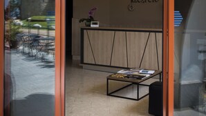 A reception area with a wooden reception desk, a black mat at the entrance, and a view of the courtyard with outdoor furniture and a building in the background. - Sándor Hotel (Pecs)