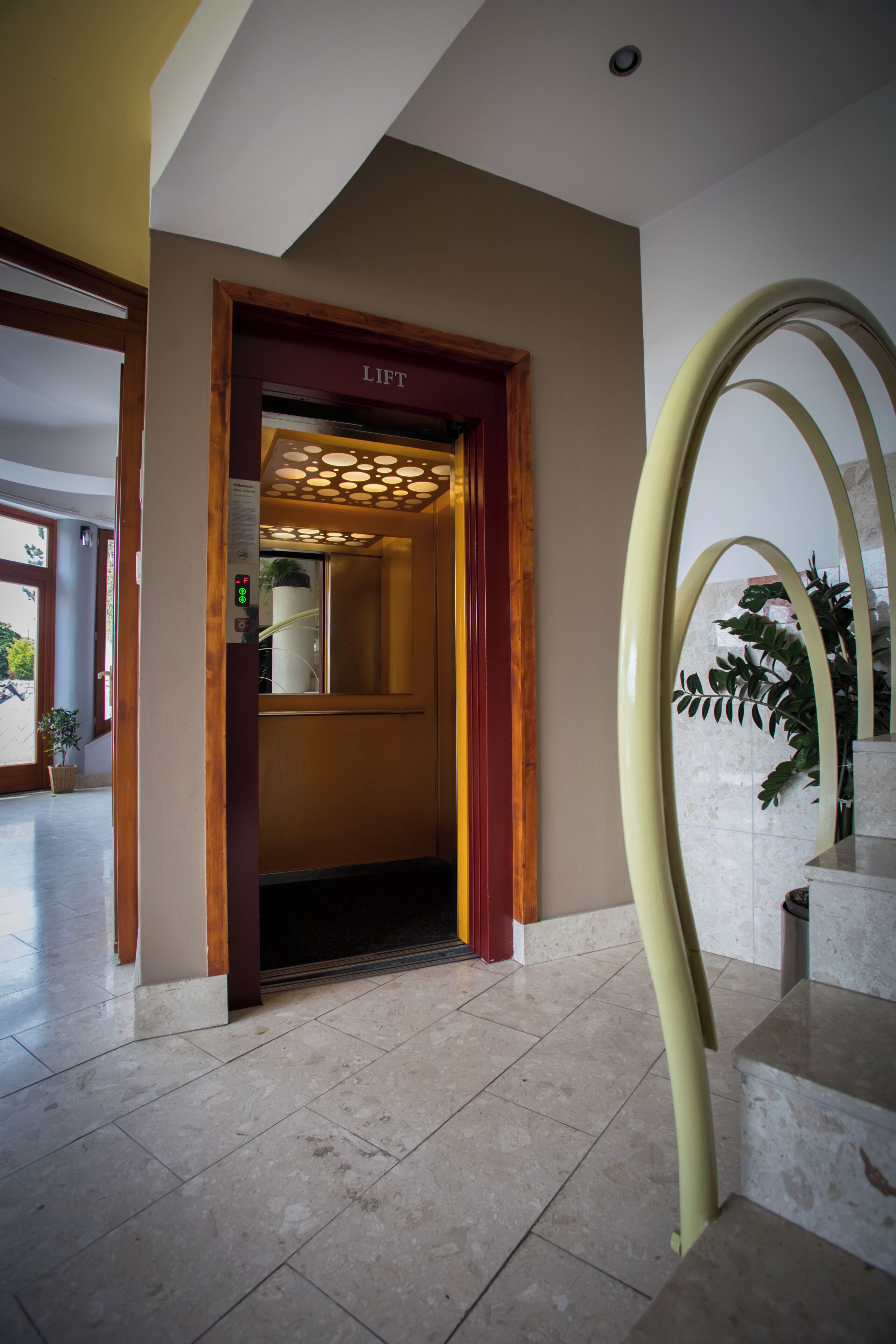 Entrada de un hotel con ascensor, una escalera de mármol y una planta en maceta.