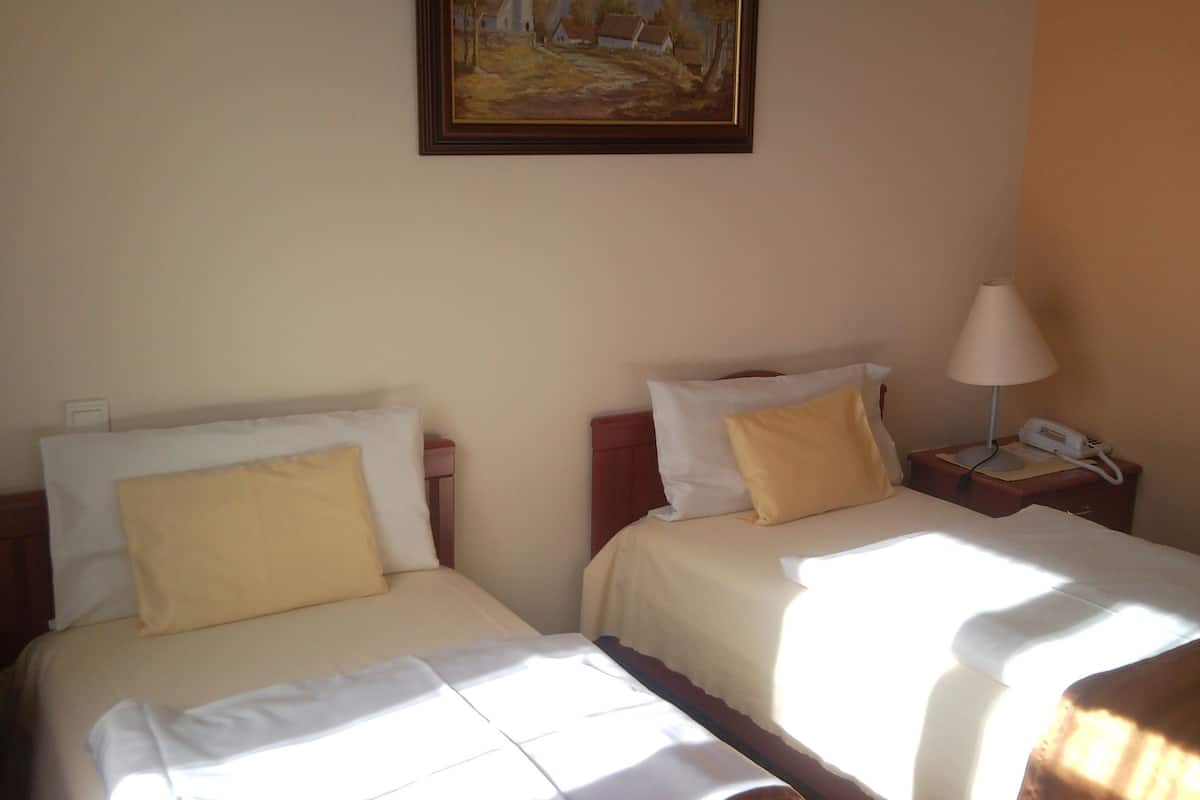 A room with two beds, each with white and yellow pillows, a bedside table with a lamp and phone, and a painting on the wall.