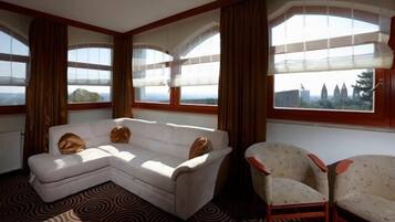 A room with a white sectional sofa and two armchairs, large windows with curtains, and a patterned carpet.