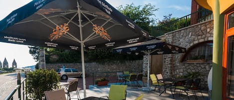 An outdoor seating area with tables and chairs, covered by umbrellas, and a stone wall in the background.