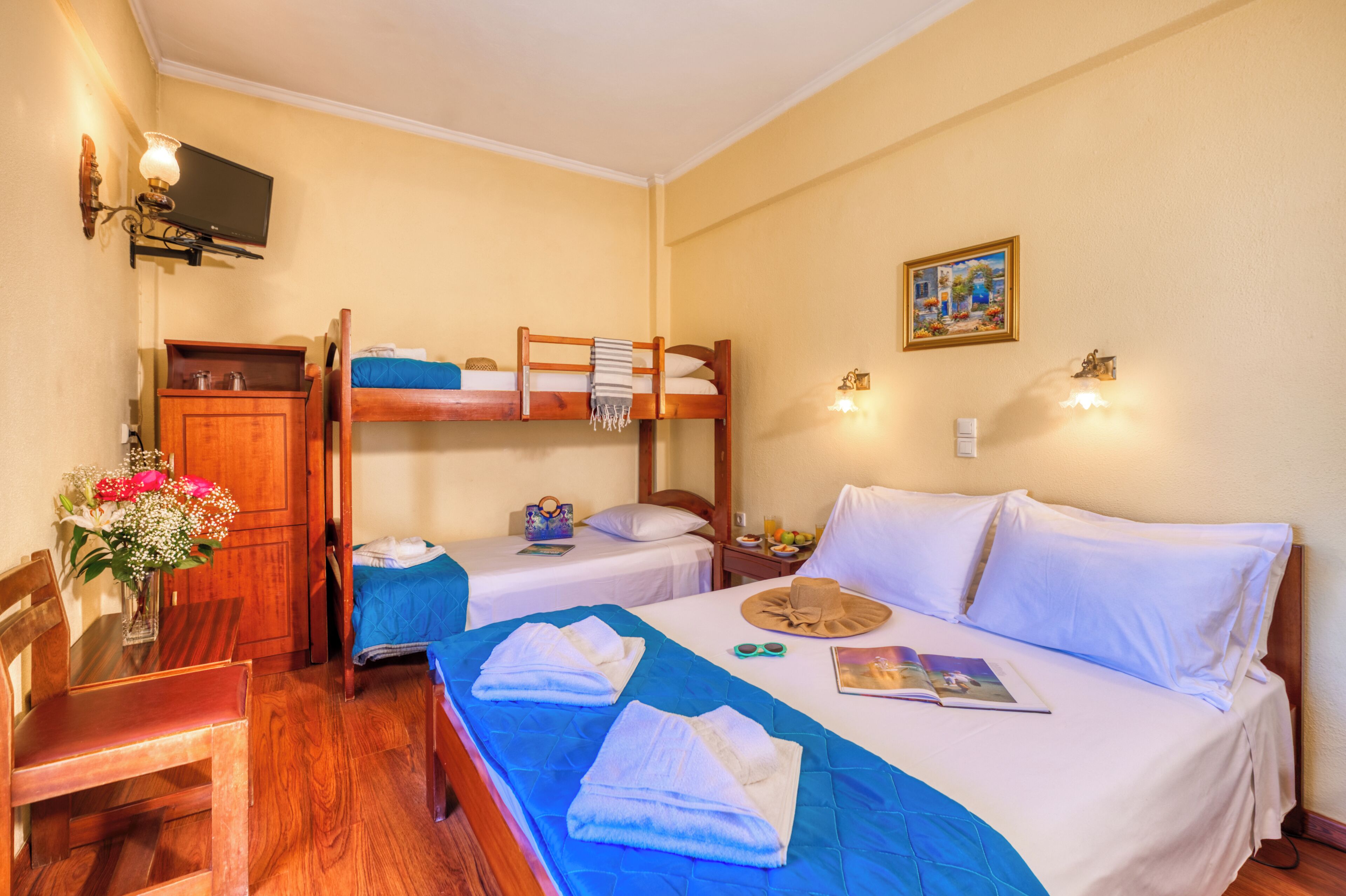 A room with two beds, a wooden desk, a chair, a small table with a vase of flowers, and a wall-mounted television.