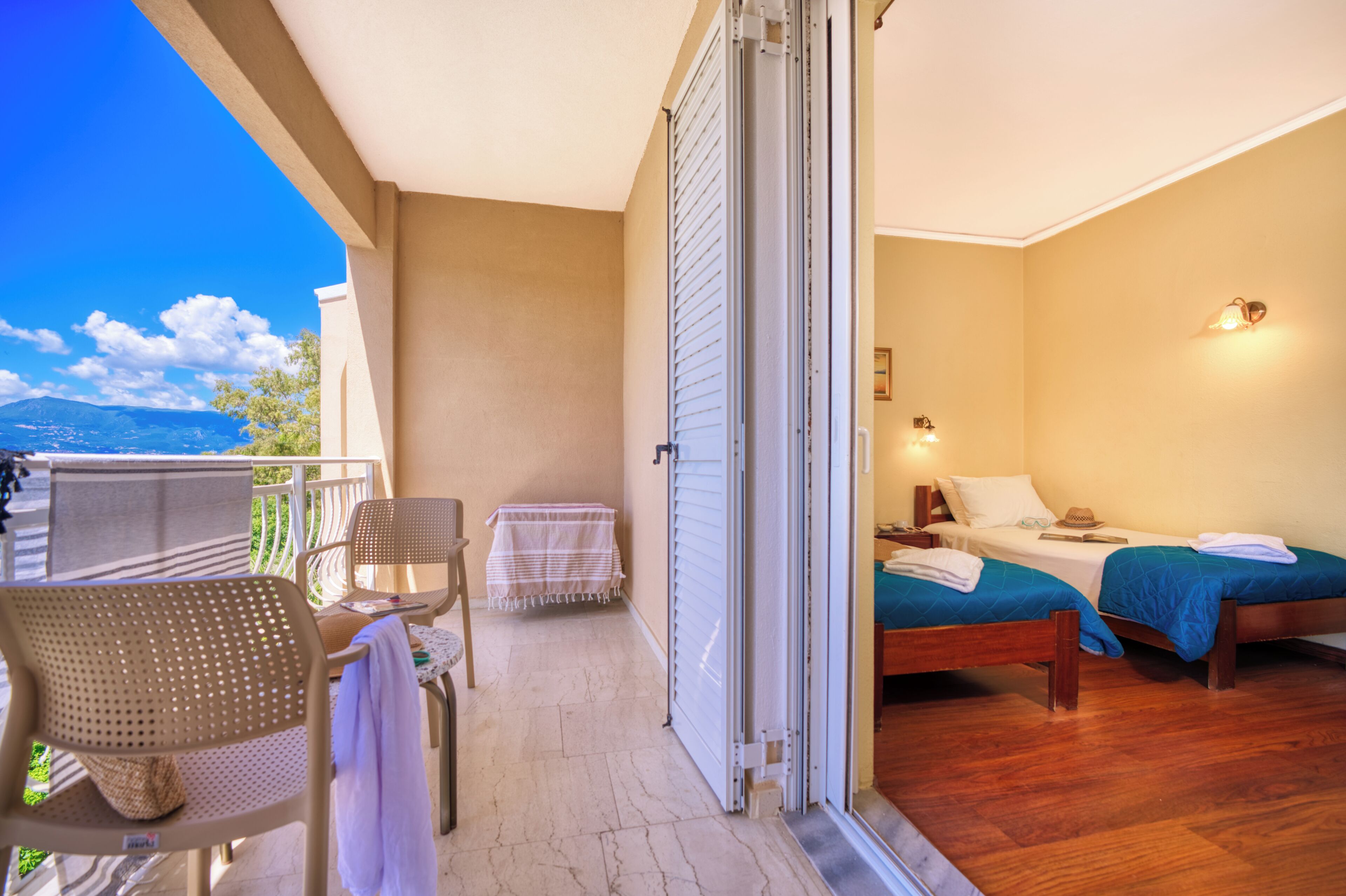 Un balcón con sillas y una mesa, una habitación con dos camas y una mesita de noche, y vistas a las montañas.
