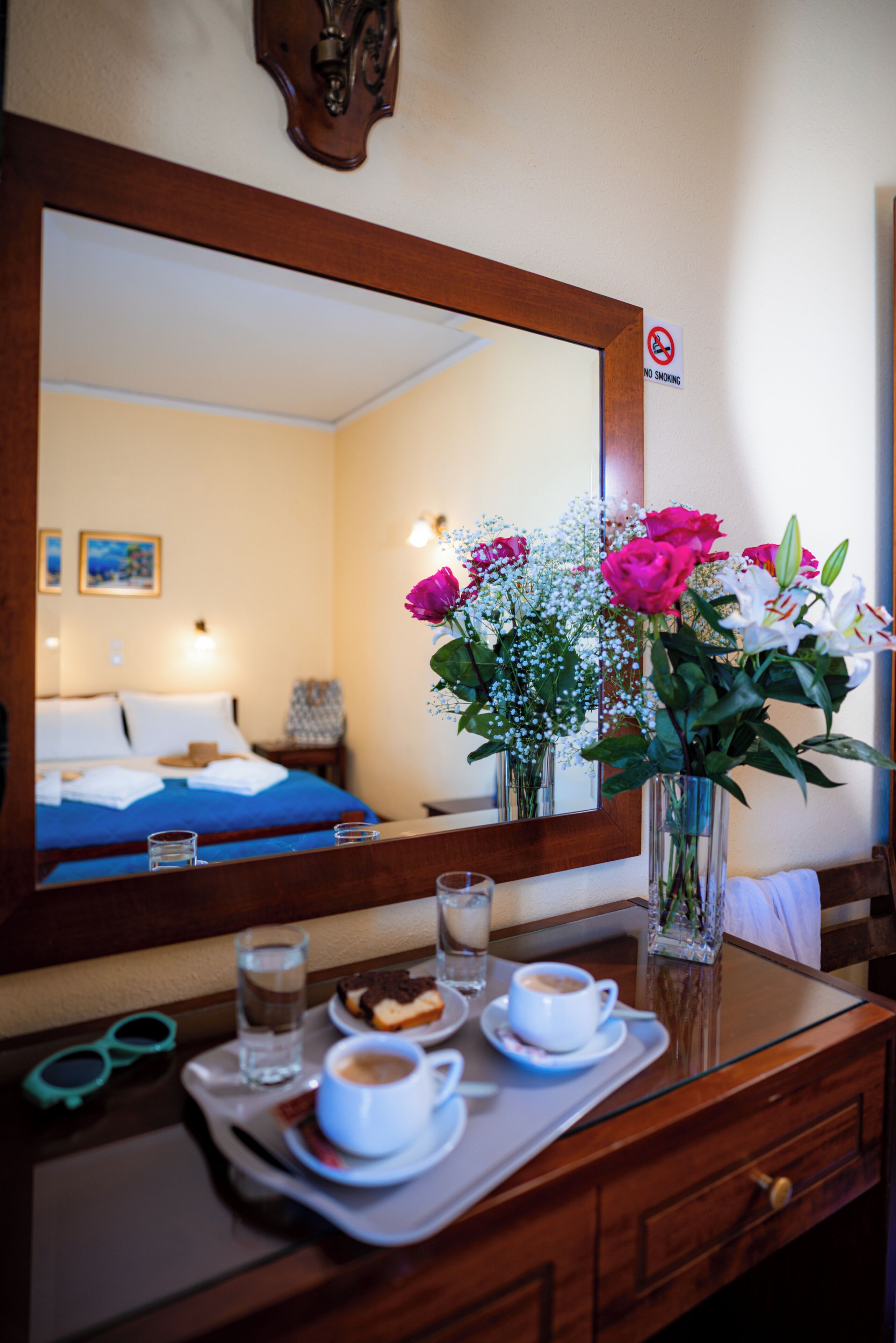 A hotel room with a bed, a mirror, a tray with a cup of coffee, a glass of water, and a vase of flowers.